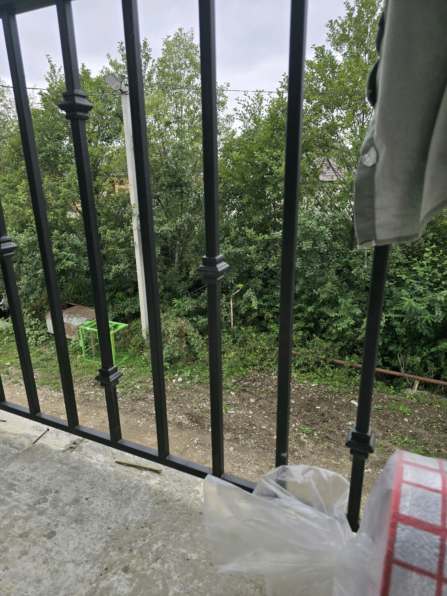 The image shows a view through a black metal railing, looking out onto a yard with lush green trees and foliage. A concrete balcony is visible in the foreground, with a roll of red and silver tape and a plastic bag on it. In the middle ground, a metal pole with a light fixture is visible amongst the trees, as is a rusty metal structure on a green frame. The ground appears to be a mix of dirt and gravel with some sparse vegetation. The sky is overcast. The location is Avtury, Russia.