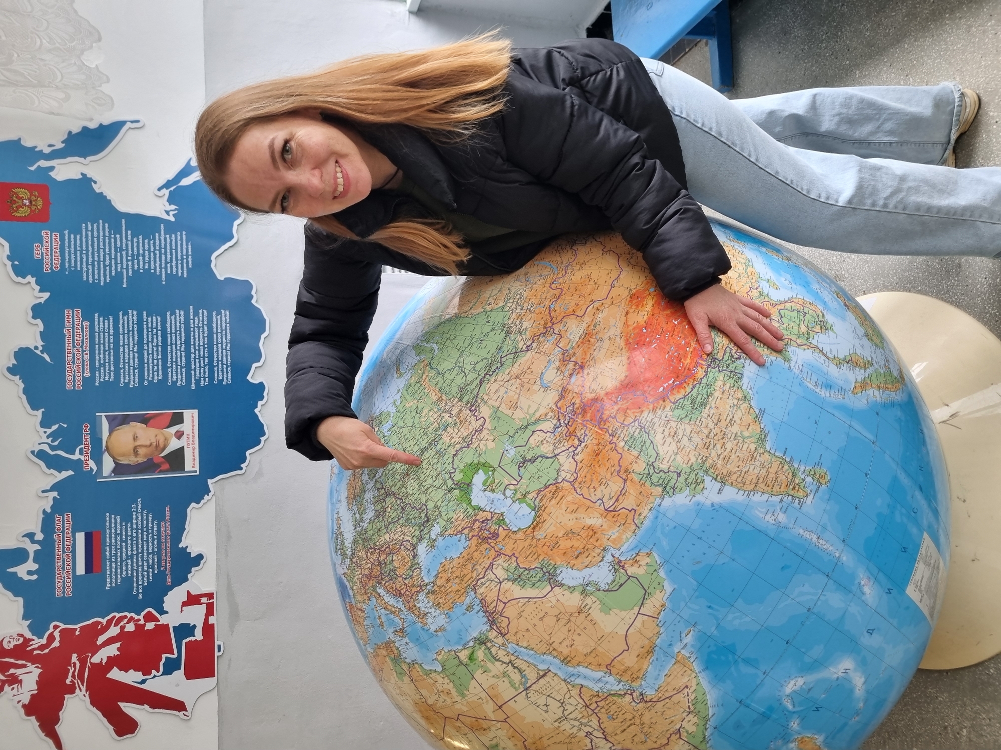 A woman is posing next to a large globe, in what appears to be an indoor location, potentially a school or public building. The woman, with light brown hair, is wearing a black puffer jacket and light blue jeans. She has her left hand on the globe, covering part of the area around Central Asia, and her right hand points towards Europe. The globe itself shows the world map, with color-coded regions.
To the left of the woman and the globe, there's a poster on the wall with information related to Russia. The poster includes the Russian coat of arms, the text of the Russian national anthem, and a picture of Vladimir Putin, identified as the President of Russia.
Based on the Russian-themed poster that includes the Russian President Vladimir Putin, the location is likely to be related to or in the city of Chaikovsky, Russia.