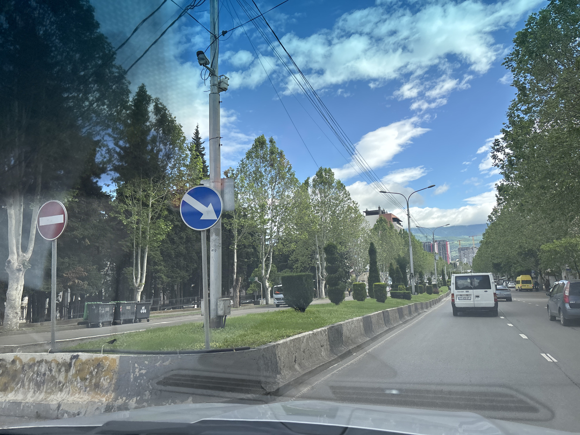 The image shows a street scene in Tbilisi, Georgia. The view is from inside a vehicle looking forward along a road. A white van is in the lane ahead. There are other vehicles further ahead. The road is lined with trees and manicured bushes, separated by concrete barriers. Two traffic signs are visible on the left side of the road: a red and white 'no entry' sign and a blue sign with a white arrow pointing left. There are utility poles with wires overhead. Buildings and mountains are visible in the distance. The sky is blue with scattered white clouds. Several trash containers are seen on the left side of the road.