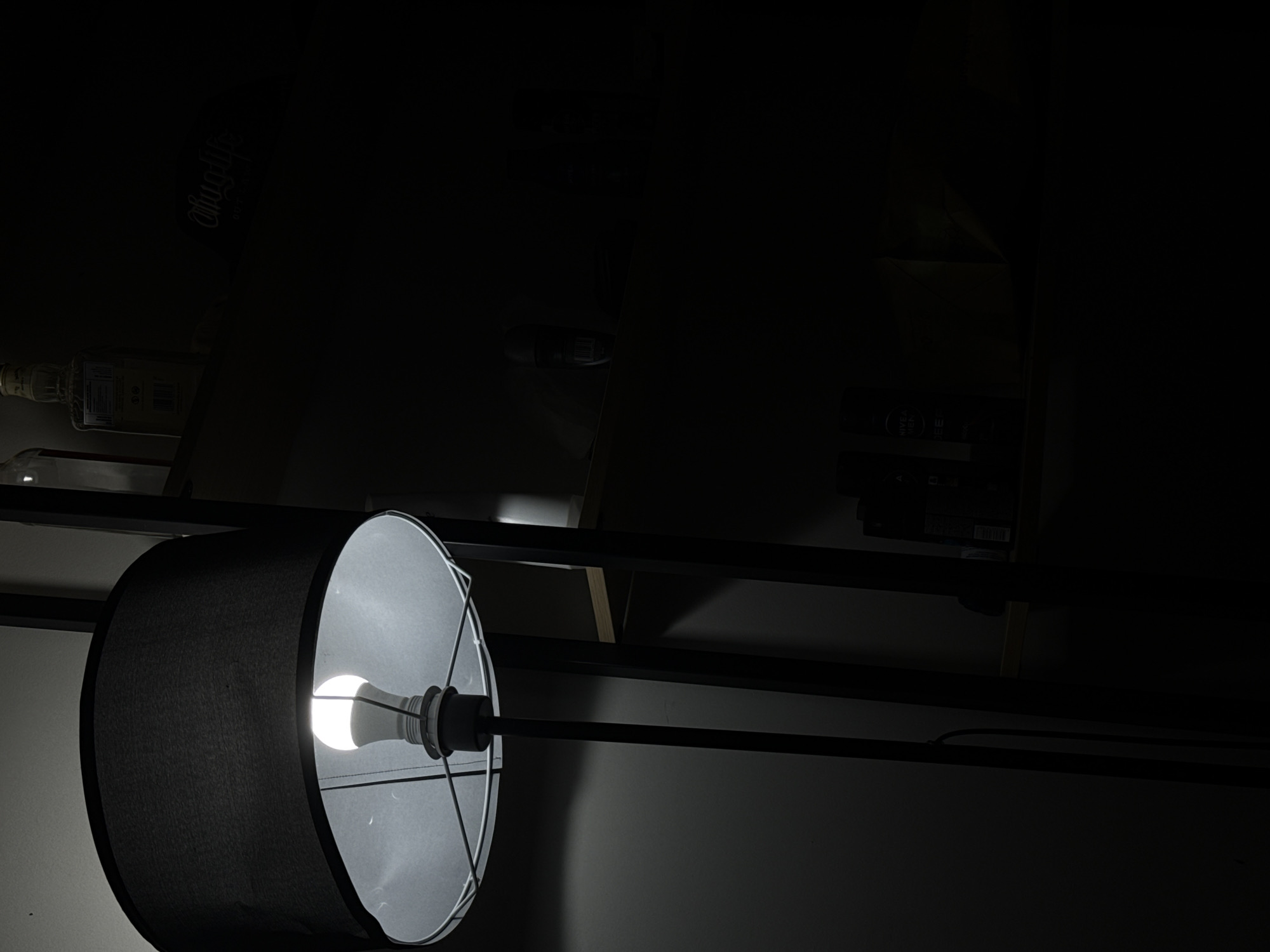 The image captures an indoor scene in Kuala Lumpur, Malaysia, characterized by low light and a prominent artificial light source. The central subject is a drum-shaped lamp shade, dark grey or black on its exterior and white on the interior, positioned on the left side of the frame and viewed from a slight angle. A bright, cool white LED light bulb is clearly visible inside the shade, emitting strong illumination that lights its immediate vicinity and casts deep shadows across the rest of the scene.
The lamp is affixed to a black metal frame, likely part of a shelving unit, with another parallel black bar running beneath it. The surrounding environment is largely shrouded in darkness. However, details emerge upon closer inspection. On a shelf in the upper left background, a glass bottle with a label is faintly visible; the label features stylized, vertically oriented text that is difficult to fully discern but resembles "Ohmygod" or a similar brand. Further into the shadows on the right, what appears to be electronic equipment or another object with reflective surfaces is vaguely outlined. In the lower right, a product box is distinguishable, bearing the clear text "NIVEA" and "MEN". The visible background elements suggest wooden or cardboard surfaces.
No people or direct activity are depicted. The scene implies a domestic or storage setting, likely captured at night or in a room with minimal natural light, relying entirely on the lamp for illumination. The strong contrast between light and shadow creates a stark, utilitarian atmosphere.