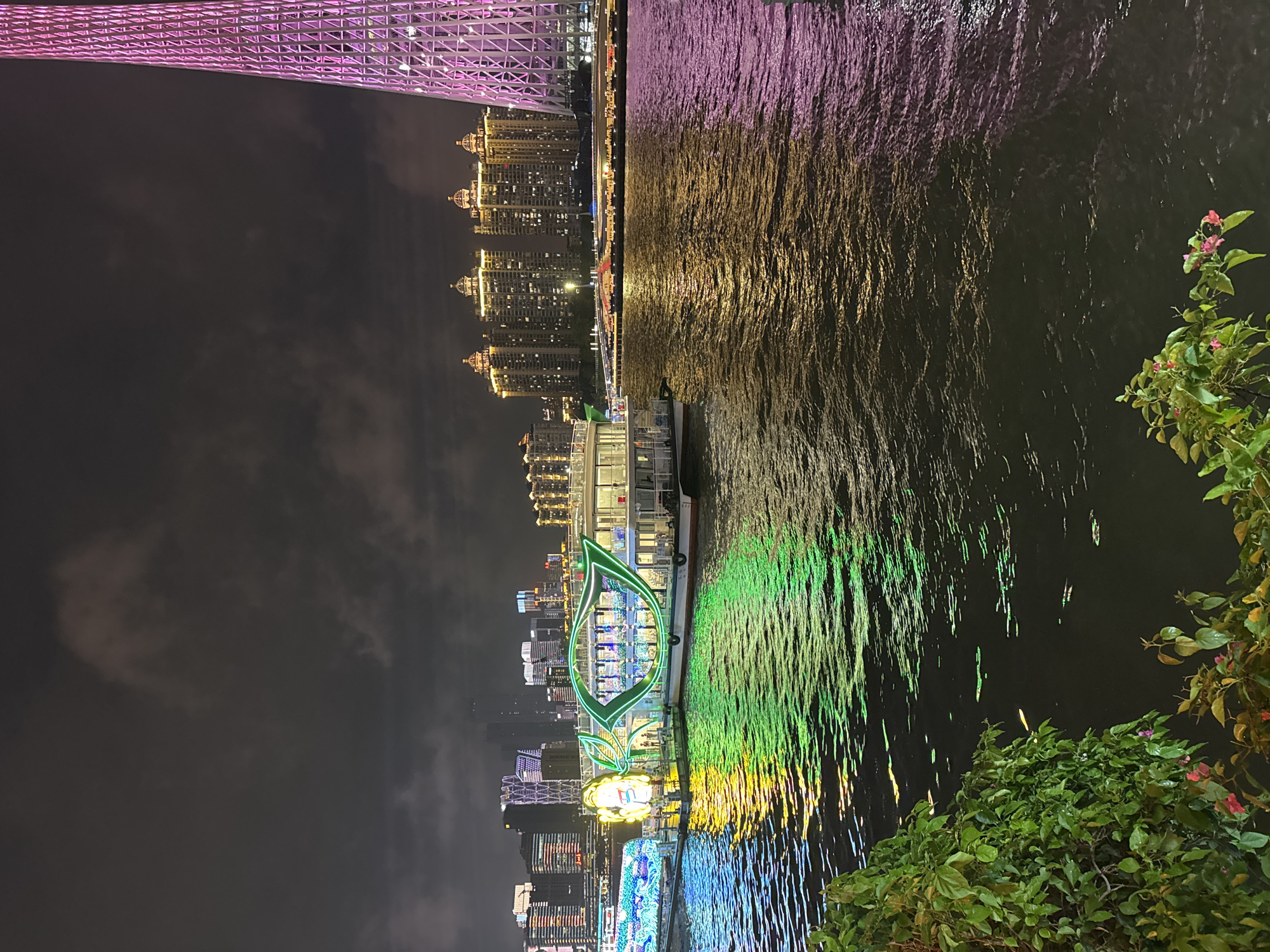 The image is a nighttime shot of a river in Tianhe District, China, with illuminated buildings and a tower reflected in the water. A brightly lit boat is docked along the riverbank. Lush greenery is visible in the foreground, partially obscuring the view. The buildings on the horizon are lit with yellow lights, while the tower glows with a pink hue. The river reflects these colors, creating a shimmering effect.