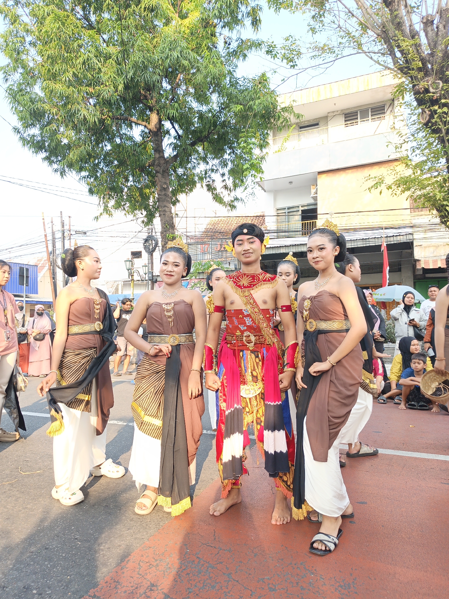 A group of four young people in traditional Indonesian attire are standing on a street in Klaten, Indonesia. Three women are dressed in brown and gold sarongs, gold necklaces, and ornate headpieces. One man in the center wears a red, gold, and black outfit with a headdress. The people are posing for a photo while a crowd watches in the background. Trees and buildings line the street, and power lines are visible overhead. The ground is paved with asphalt and marked with a red painted area.