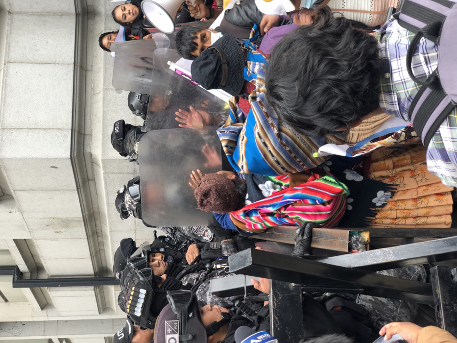 A group of people face a line of riot police behind shields. The crowd includes individuals wearing traditional clothing and carrying a megaphone. Some people have their hands pressed against the shields. The riot police are wearing full gear, including helmets, and some have visible equipment on their vests. The scene appears to be a protest or demonstration in front of a building.