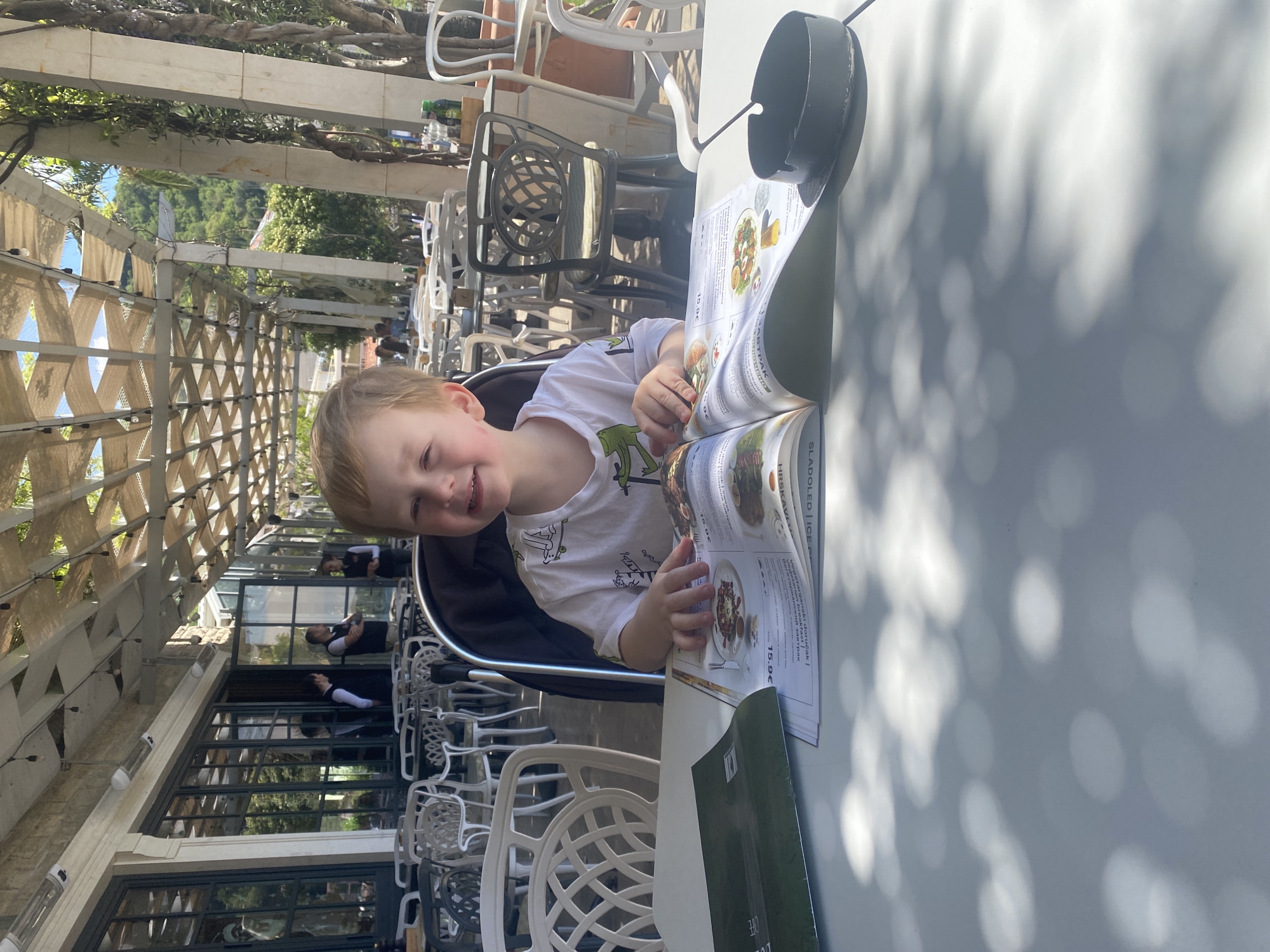 A young boy sits at a table in an outdoor restaurant in Bečići, Montenegro. The restaurant has a lattice-covered awning providing shade. The boy, wearing a white t-shirt with a green dinosaur design, looks at a menu, his hand on the page. A black container sits on the table beside the menu. The table surface is light-colored, and dappled sunlight casts shadows. Behind the boy are several empty white chairs, and in the background, glass windows reflect the image of servers.