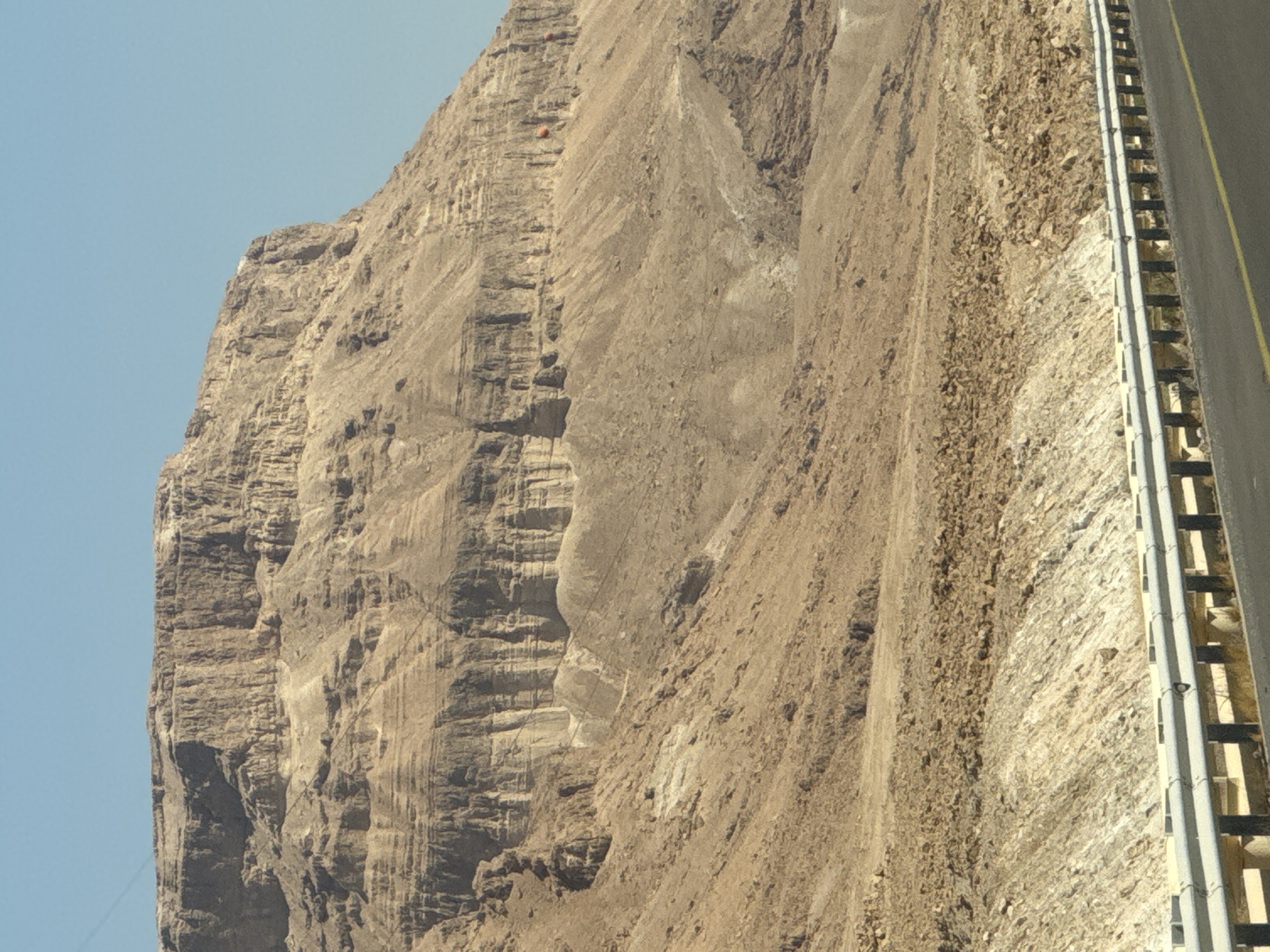 The image depicts a vast, arid mountainous landscape under a clear blue sky. A rugged, layered cliff face dominates the upper portion of the frame, exhibiting erosion and geological stratification. The lower slopes are covered in loose scree and sparse vegetation. In the foreground, a paved road runs horizontally, bordered by a metal guardrail. The scene appears to be in the Tamar Regional Council, Israel, likely a desert environment like the Judean Desert, given the arid conditions and rock formations characteristic of the region. There are no people or discernable animals visible in the image. Several orange objects are visible on the cliff face, possibly paragliders or large debris.