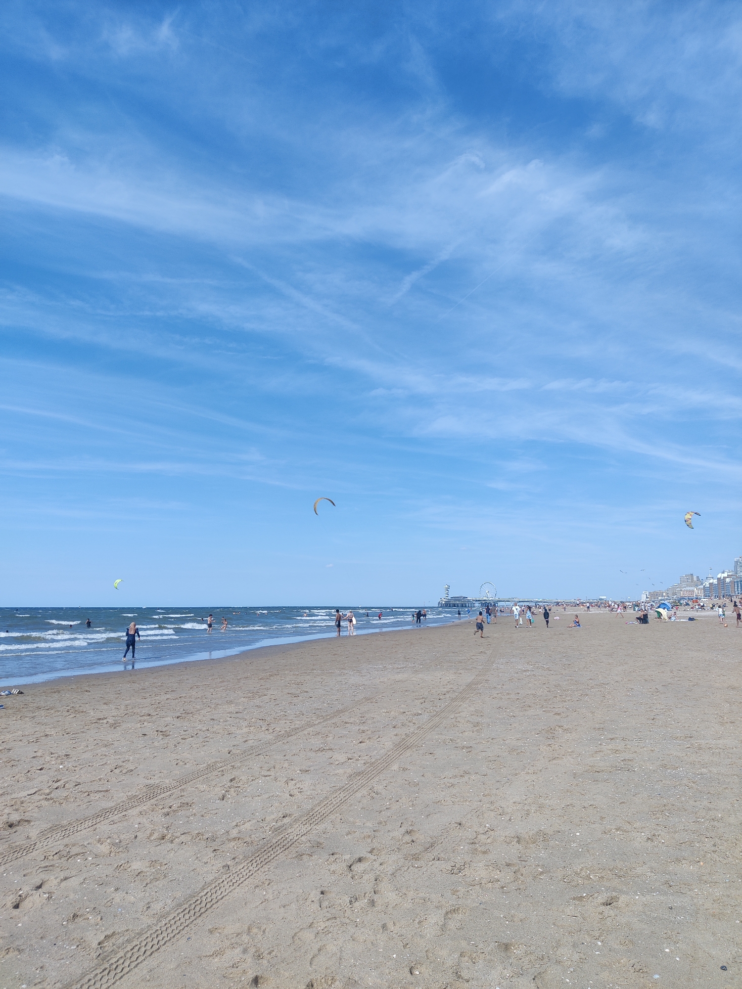 The image depicts a wide sandy beach on a bright, clear day with a blue sky scattered with wispy clouds. In the foreground, tire tracks are visible in the sand, suggesting recent vehicle passage. The beach is populated with numerous people engaged in various activities. Some individuals are walking along the shore, while others are in the water, swimming or playing in the waves. Two kitesurfers are visible in the ocean, one with a yellow and brown kite, and another with a lime green kite. In the distance, a pier extends into the sea, topped with a Ferris wheel and other structures, indicating an amusement area. The shoreline is lined with buildings, suggesting an urban environment adjacent to the beach. The scene captures a lively atmosphere at Scheveningen beach, a popular seaside resort in The Hague, Netherlands.