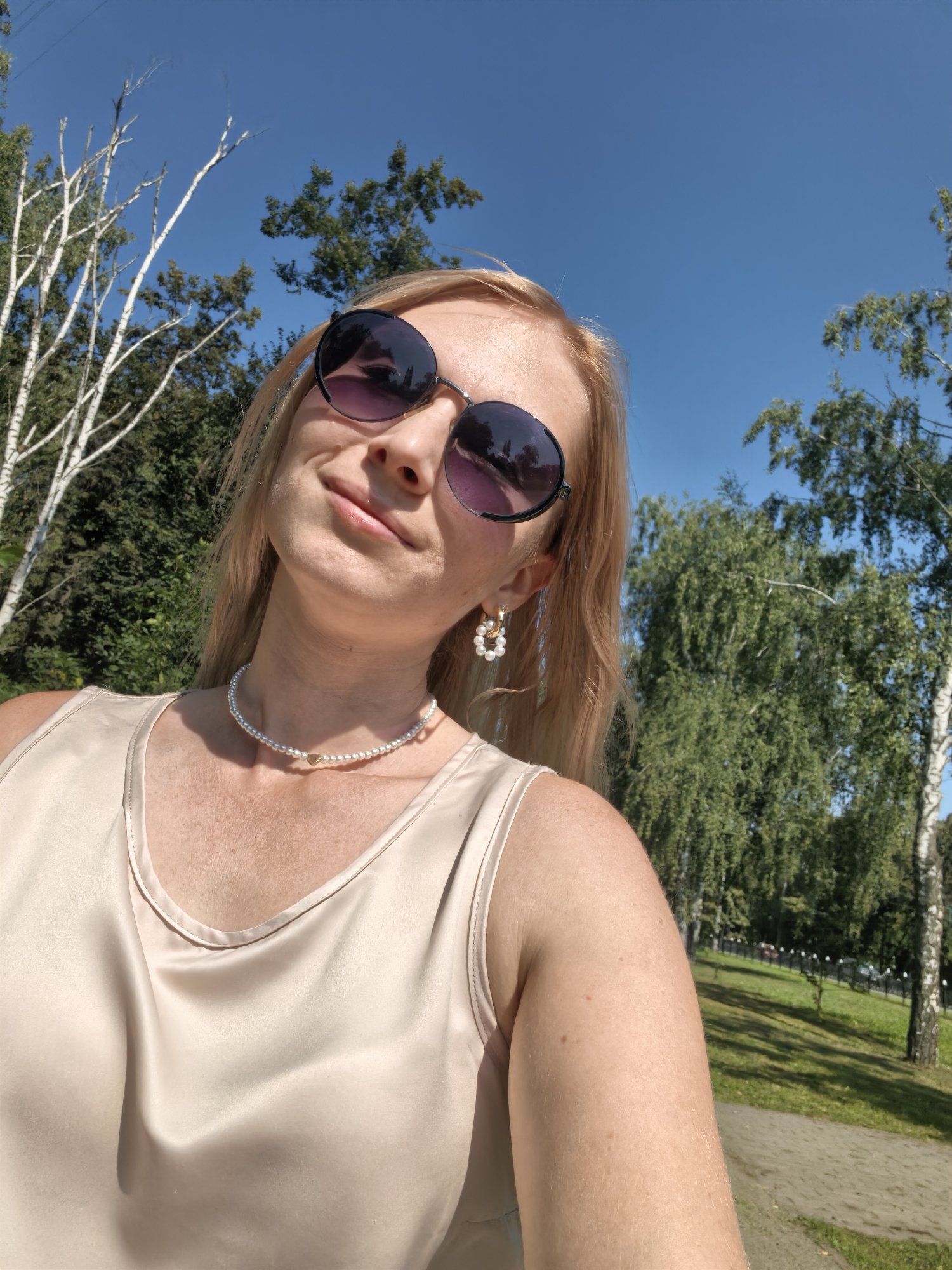 A woman with blonde hair, wearing sunglasses, a beige top, a pearl necklace, and pearl earrings, poses for a selfie outdoors in Sterlitamak, Russia. The background consists of trees and a clear blue sky. The woman is looking up and smiling. The image appears to be taken in a park or a grassy area.