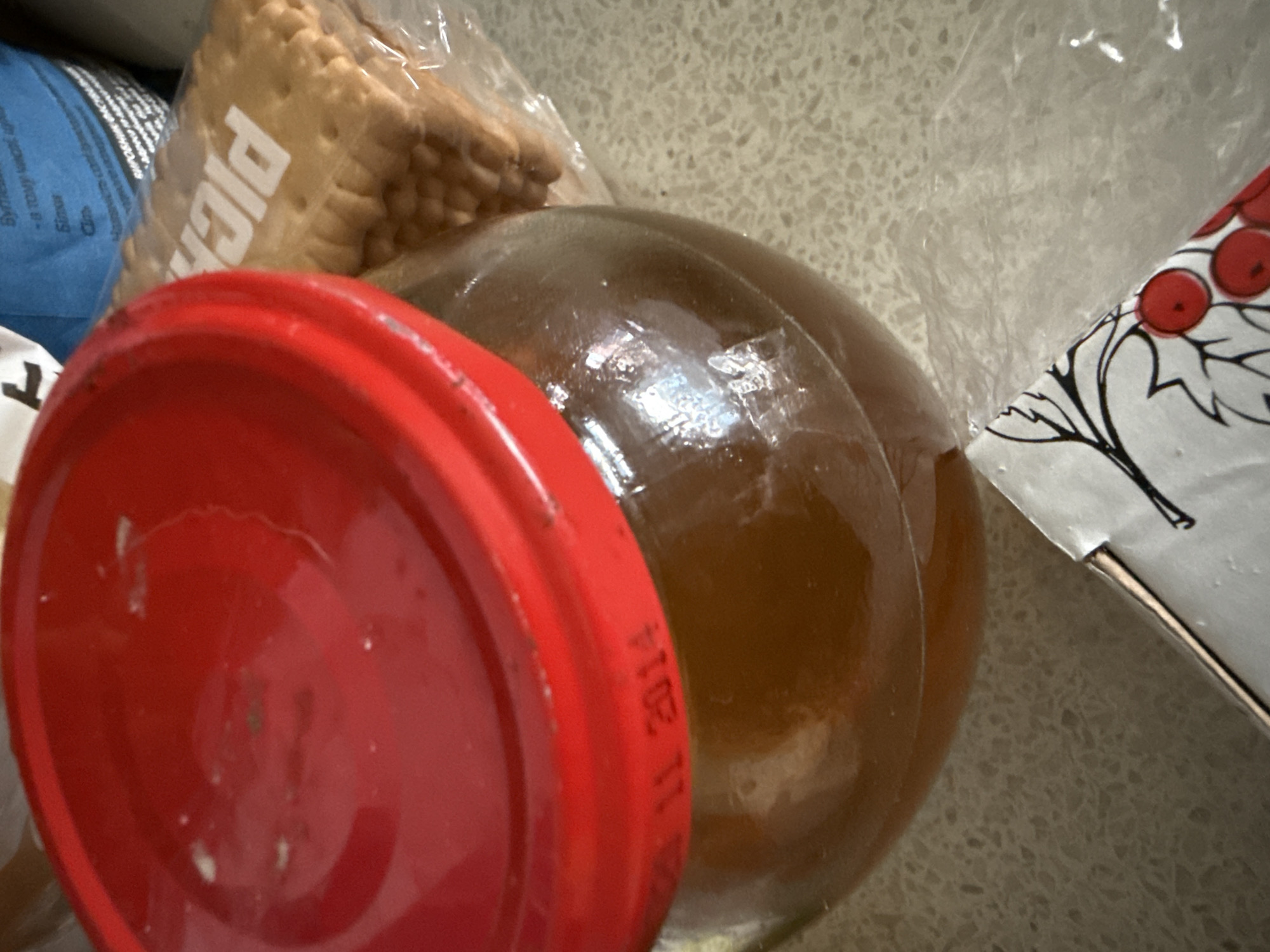 The image depicts a close-up, overhead shot of a jar of honey with a red lid. The lid has some white specks on it. Embossed on the lid's edge, or possibly on the glass jar beneath it, is text that reads "24 11 2014". To the right of the honey jar is a stack of rectangular crackers in clear plastic packaging. The crackers are golden brown with a grid pattern on their surface and have the word "PIC" visible on them. In the background, a portion of a blue and white package is visible, with some text in what appears to be Cyrillic script. The overall setting appears to be a kitchen counter, characterized by a speckled surface. The image is taken in Okhtyrka, Ukraine.