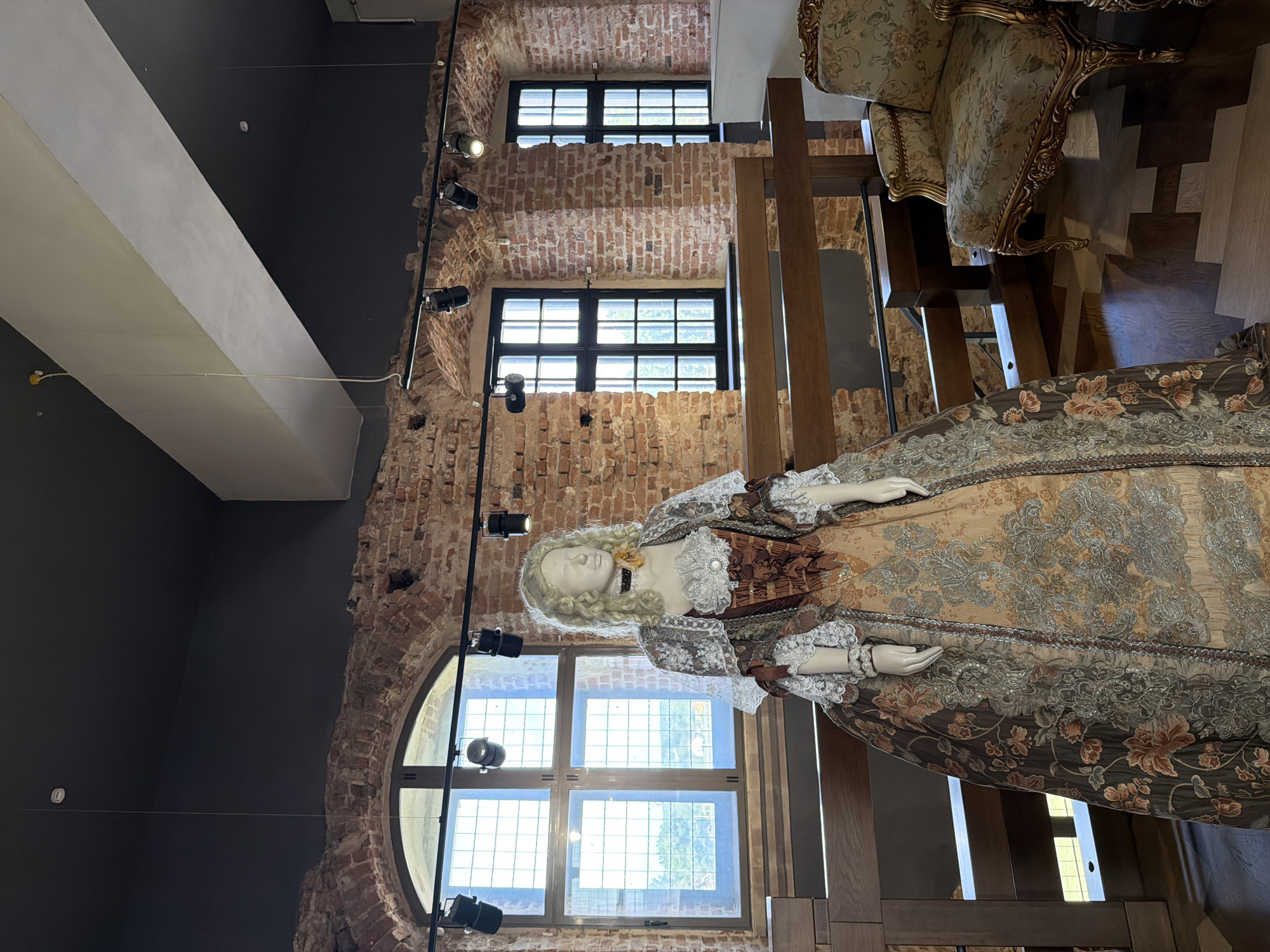 This media file displays a mannequin dressed in a lavish, ornate historical gown, positioned in what appears to be a museum or exhibition space. The setting features exposed brick walls and large, multi-paned windows that allow natural light to illuminate the scene. Modern track lighting is visible, casting spotlights on the display. To the right, a richly upholstered armchair with intricate golden detailing suggests a historical or opulent theme for the exhibit. The mannequin itself is adorned with a pale blonde wig, and its outfit is characterized by intricate floral patterns, lace trim, and layered fabrics in shades of brown, gold, and grey. The gown has a wide skirt and elaborate sleeves, typical of 18th-century fashion. The overall impression is one of historical display and preserved artistry. There is no visible text within the image. The time of day appears to be daytime, with light filtering through the windows.