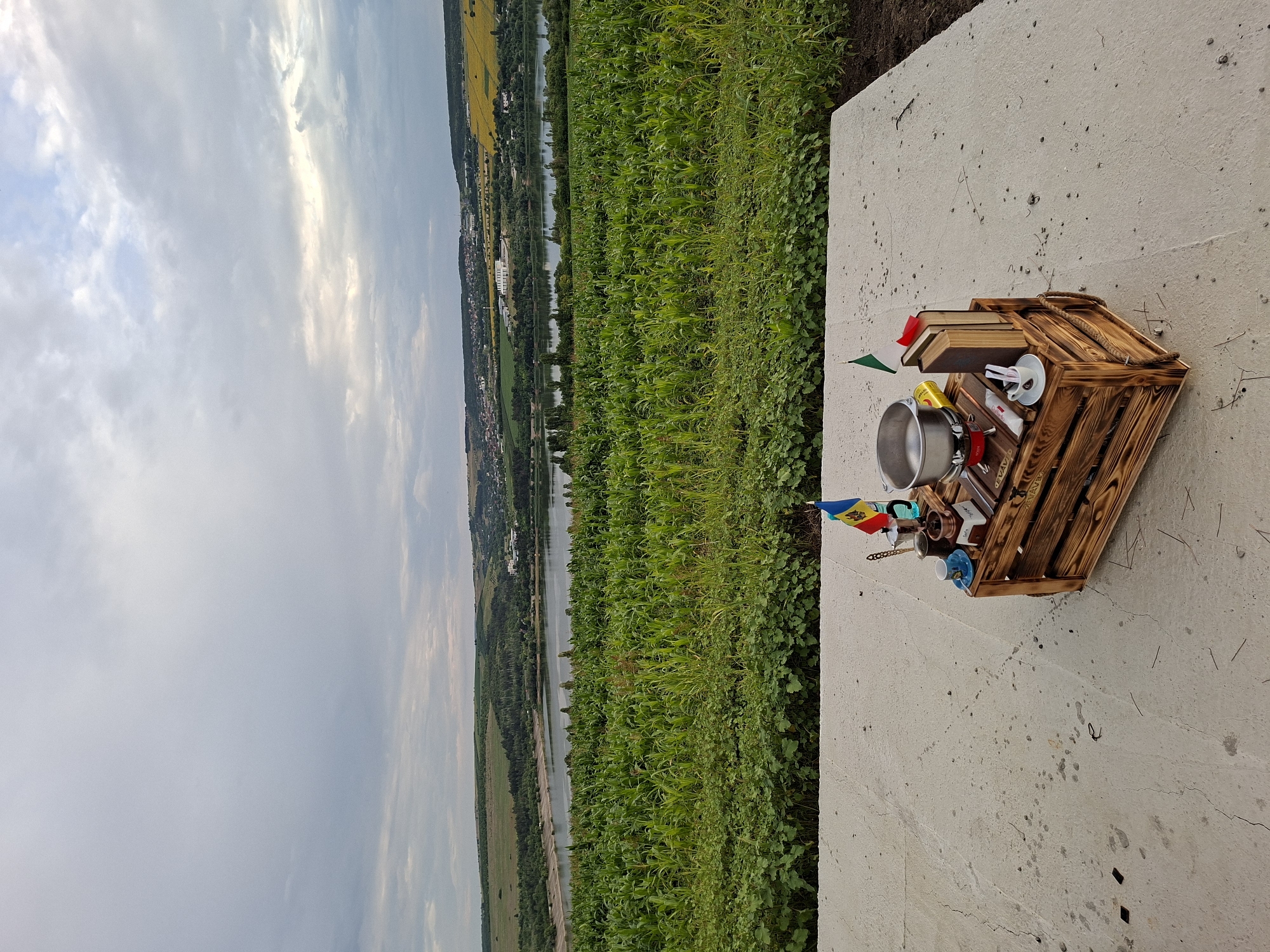 The image depicts an outdoor scene overlooking a landscape that appears to be Sociteni, Moldova. The foreground features a weathered wooden crate placed on a concrete platform. The crate is adorned with various items, including a small portable stove with a metal pot, a coffee grinder, a small blue cup and saucer, two books, a yellow can, and two small flags, one of which resembles the flag of Moldova and the other the flag of Italy. The background reveals a lush green field, likely a cornfield, leading to a body of water, possibly a lake or river, with a town or village situated on the opposite bank. Rolling hills, some dotted with trees and houses, form the horizon under a cloudy sky. The overall atmosphere is serene and rural.
