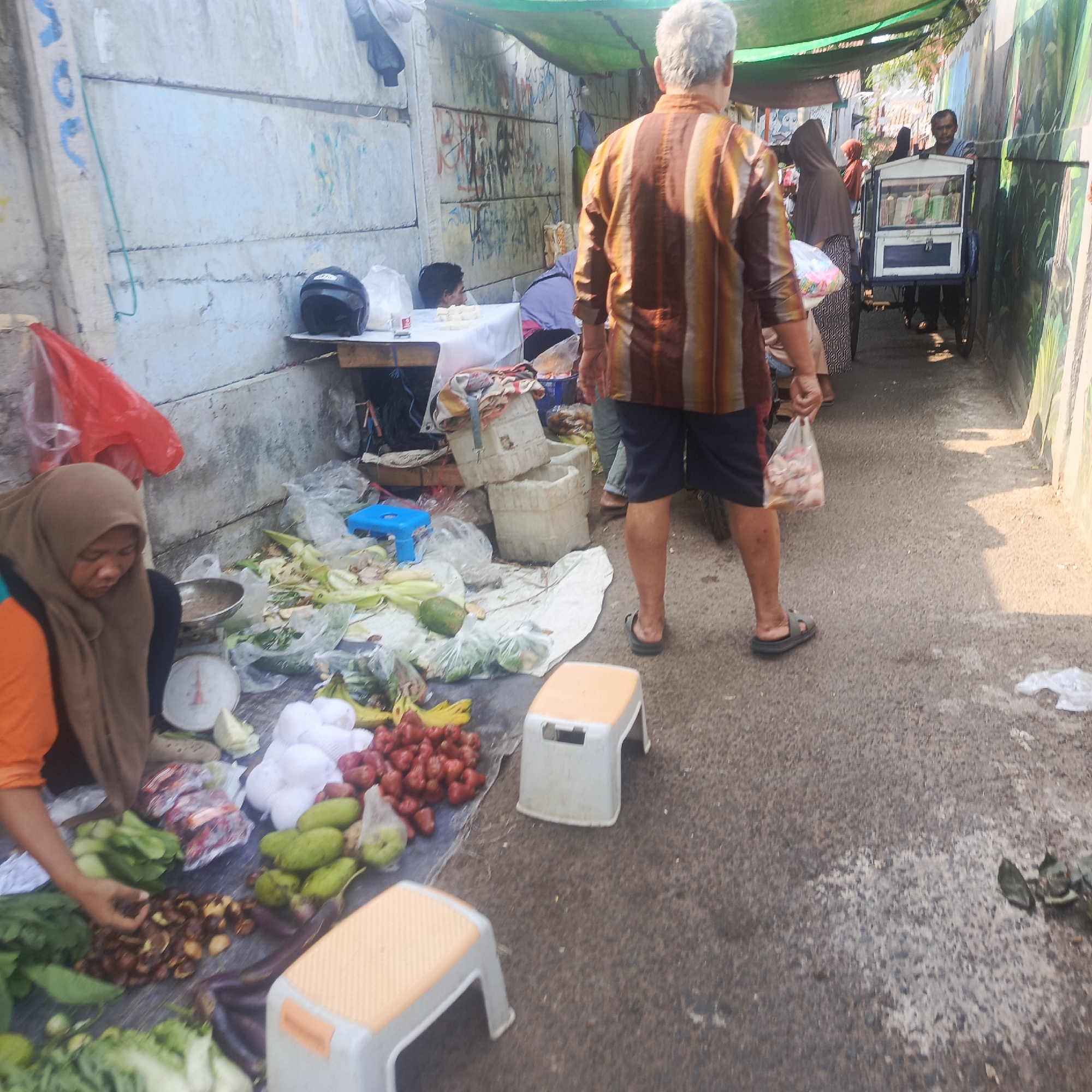 The image depicts a street scene in South Jakarta, Indonesia, likely a market or roadside stall. In the foreground, a woman wearing a brown hijab and an orange shirt is seated at a table with a scale, surrounded by various fruits and vegetables like red apples, green mangoes, white fruits, and leafy greens. To her left, a red plastic bag hangs. Behind her, a man with gray hair and a striped shirt walks away from the camera, carrying a plastic bag. In the background, other people and stalls are visible, including a vendor with a cart and a person in a purple hijab. The environment appears to be an outdoor setting with a concrete wall covered in graffiti on the left and a green tarp providing shade overhead. The ground is asphalt.