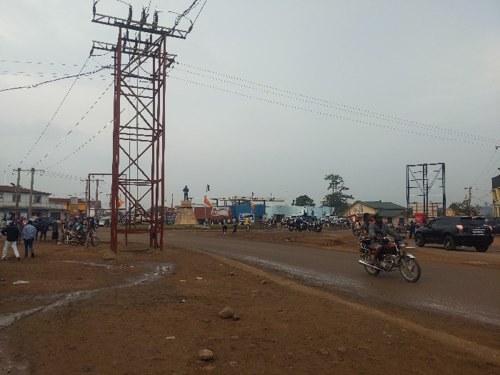 The image captures an unpaved urban intersection in Beni, Democratic Republic of the Congo, under an overcast sky. The foreground consists of reddish-brown dirt interspersed with muddy puddles and small rocks. A prominent, tall, red lattice-work power transmission tower stands left of center, supporting multiple overhead power lines that extend across the scene.
In the midground, a dirt road runs diagonally from left to right. A silver motorcycle with two occupants, both wearing dark clothing, travels rightward on this road. To its right, a dark-colored SUV is visible. On the left side of the road, approximately two dozen individuals are standing or walking, some in proximity to several parked motorcycles. Further into the scene, at what appears to be a central roundabout, a raised monument supports a blue-clad statue.
The background features various one and two-story buildings, some with visible storefronts, lining the perimeter of the intersection. Additional utility poles and power lines are present, crisscrossing the sky. On the far right, a large, bare metal framework, consistent with a billboard structure, stands against the horizon. The scene depicts active daily urban life.