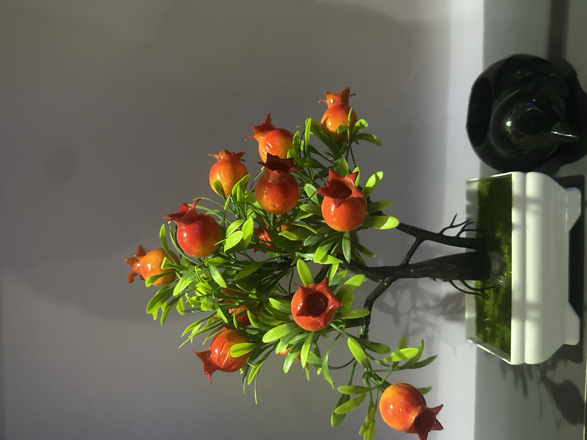 The media file shows a close-up, vertically oriented shot of an artificial pomegranate tree with bright orange and red fruits and green leaves. The tree is rooted in a white planter filled with artificial green moss. To the right of the planter is a black, glossy object, possibly a decorative piece or a part of a larger item. The background is a plain, light-colored wall, suggesting an indoor setting. The lighting appears to be artificial, creating shadows on the wall behind the tree. The overall scene is still and decorative, with no people or discernible activity. There is no visible text in the image. The image was likely taken indoors, possibly in a home or a commercial space.