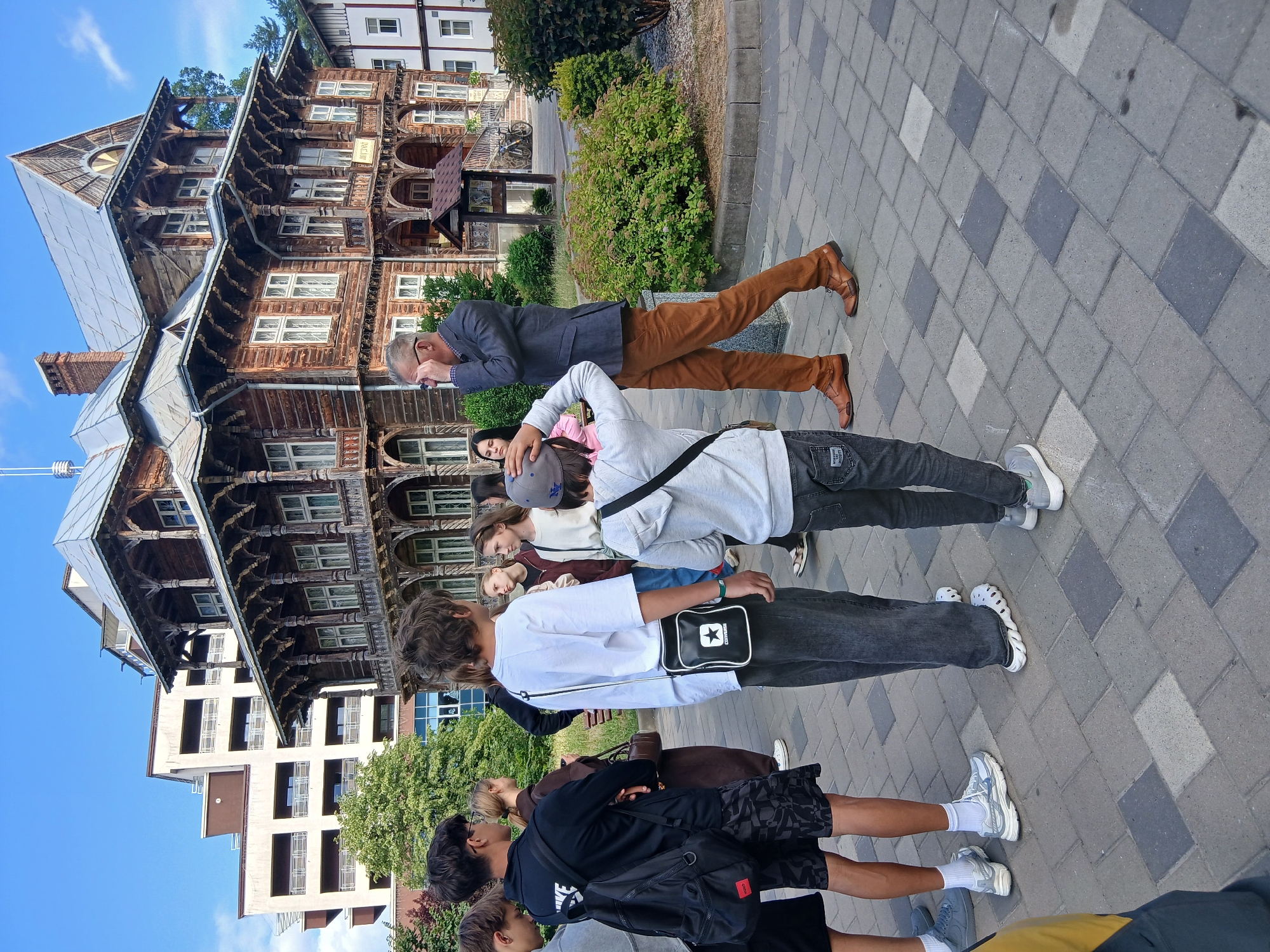 The image depicts a group of people standing on a paved area in front of an old wooden building with intricate architectural details. A man in a suit and brown trousers is addressing the group, with his hand on his face as if adjusting his glasses. Several young people, appearing to be students, are listening attentively. The wooden building has a sign that appears to read "MUSEUM". In the background, a modern multi-story building is visible. The environment suggests an outdoor setting in a city, with trees and shrubs present. The overall scene is a guided tour or educational visit.