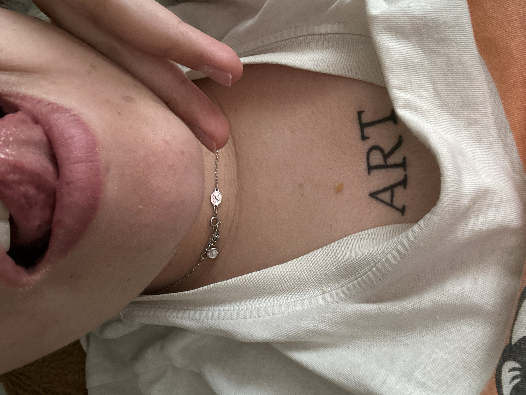 A close-up, angled shot shows a person's face and upper body. The person has their tongue extended. A finger is near the person's neck, which is adorned with a silver chain necklace. A tattoo of the word "ART" is visible on the chest, near the neckline of a white t-shirt. The setting or location within Sofia, Bulgaria, is not discernible from the image.