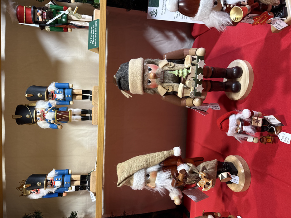 The image displays several wooden nutcrackers, likely for sale in a store. On the left, three traditional blue and black nutcrackers are displayed on a wooden shelf, alongside a sign noting, "To Purchase Please Ask An Associate for Help." To the right, on a red surface, are three nutcrackers with a folk art or woodland theme. One is in a brown hat, another in a tan cap with a burlap sack, and the third in a red Santa hat. Price tags, including numbers "101081-6" and "101151-1" and costs like "104.00," are attached to the figures. A sign with "Over 300 years ago wood turning" is also visible.