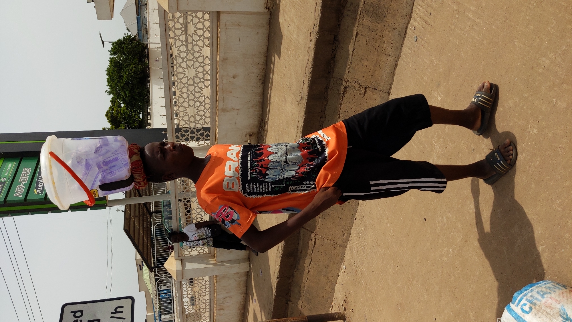 A young person stands outdoors, likely on a street or roadside in Ado, Nigeria, engaged in the activity of carrying goods for sale. The individual, dressed in an orange t-shirt, black shorts/capris with white stripes, and dark slides (sandals) with "adida" visible, balances a translucent white plastic bucket on their head. The bucket contains multiple clear plastic sachets of water, one clearly labeled "Queen Water".
The scene is brightly lit by the sun, casting strong shadows on the ground, suggesting it is mid-day. In the background, there's a green sign partially visible with text like "NR-GAS", "Autocare", and "AutoM", indicating a petrol station. To the right, a decorative white wall or fence with intricate geometric patterns is present. Another white sign is partially visible, showing "ed", "a", and "/h".
The orange t-shirt of the main subject features a graphic design and text, including "BRA" (likely part of "BRAND"), "Authorised Studio", "CRA", "BRAC", and three stars (★★★). On the ground in the bottom right, a white sack or bag is visible with "CRA" and "BRAC" printed on it. The individual maintains a neutral expression, focused on balancing the load. The overall setting depicts a common street vending scene in a warm climate.