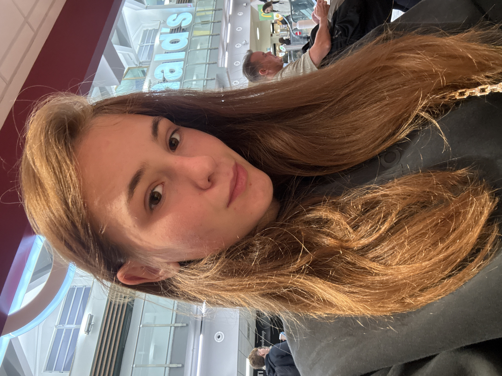 A young woman with long, reddish-brown hair is taking a selfie in an indoor environment, likely an airport or shopping mall, as indicated by the signage for "McDonald's" visible in the background. She is wearing a dark sweatshirt. Behind her, other people are visible, including a man seated at a table looking at a device and a woman standing near an escalator. The lighting suggests an overhead, artificial source. The location is not Pisa, Italy.