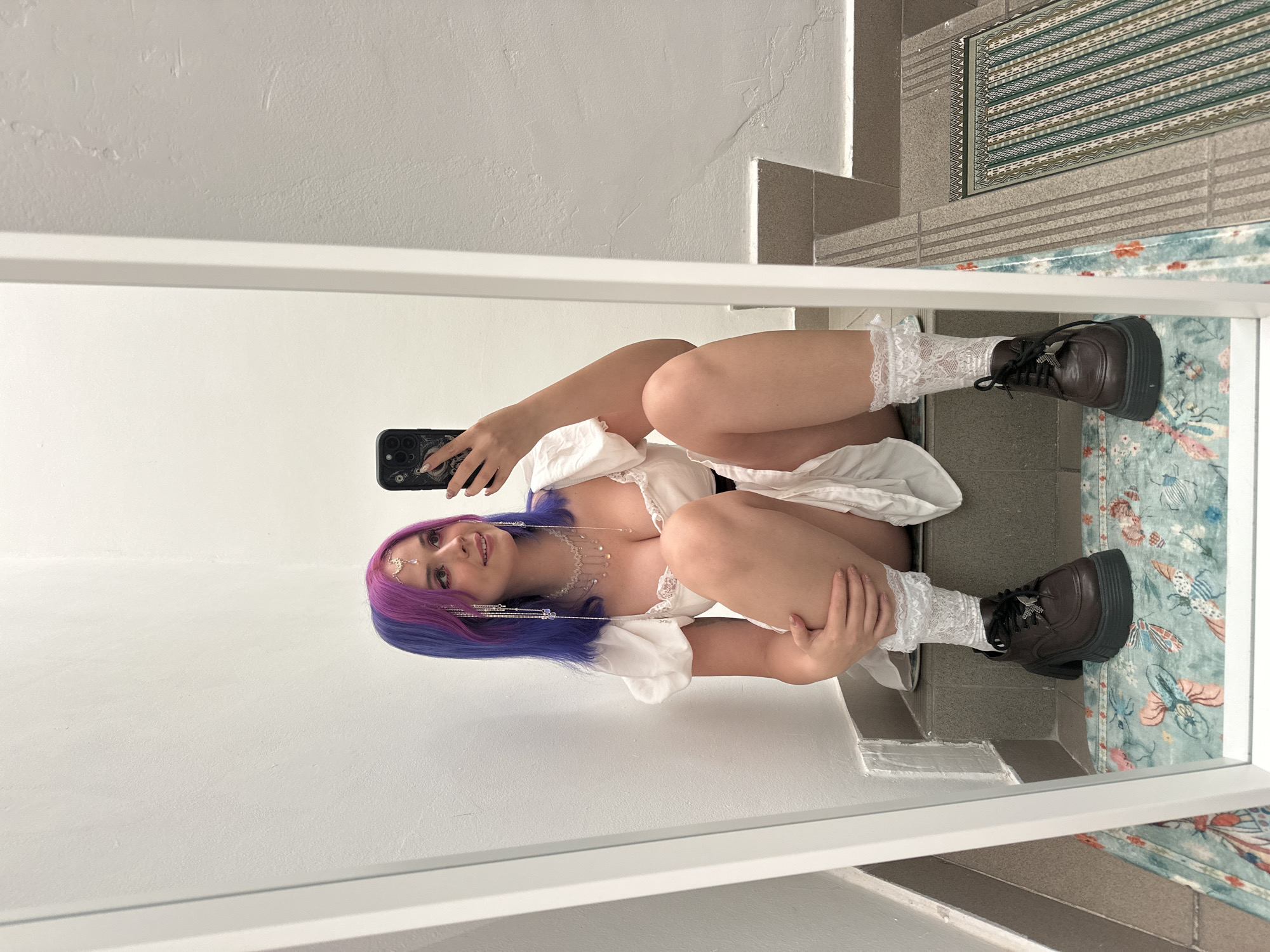 The image shows a reflection of a person in a mirror. The person has blue and purple hair and is wearing a white dress, white lace socks, and dark brown platform shoes. They are holding a phone and taking a picture of themselves. The background behind the person is a white wall and a staircase with a patterned rug.
While the city of Zagreb, Croatia is mentioned, there is no information in the image to confirm that the location is in this city. Therefore, this part of the prompt cannot be verified from the visual information provided.