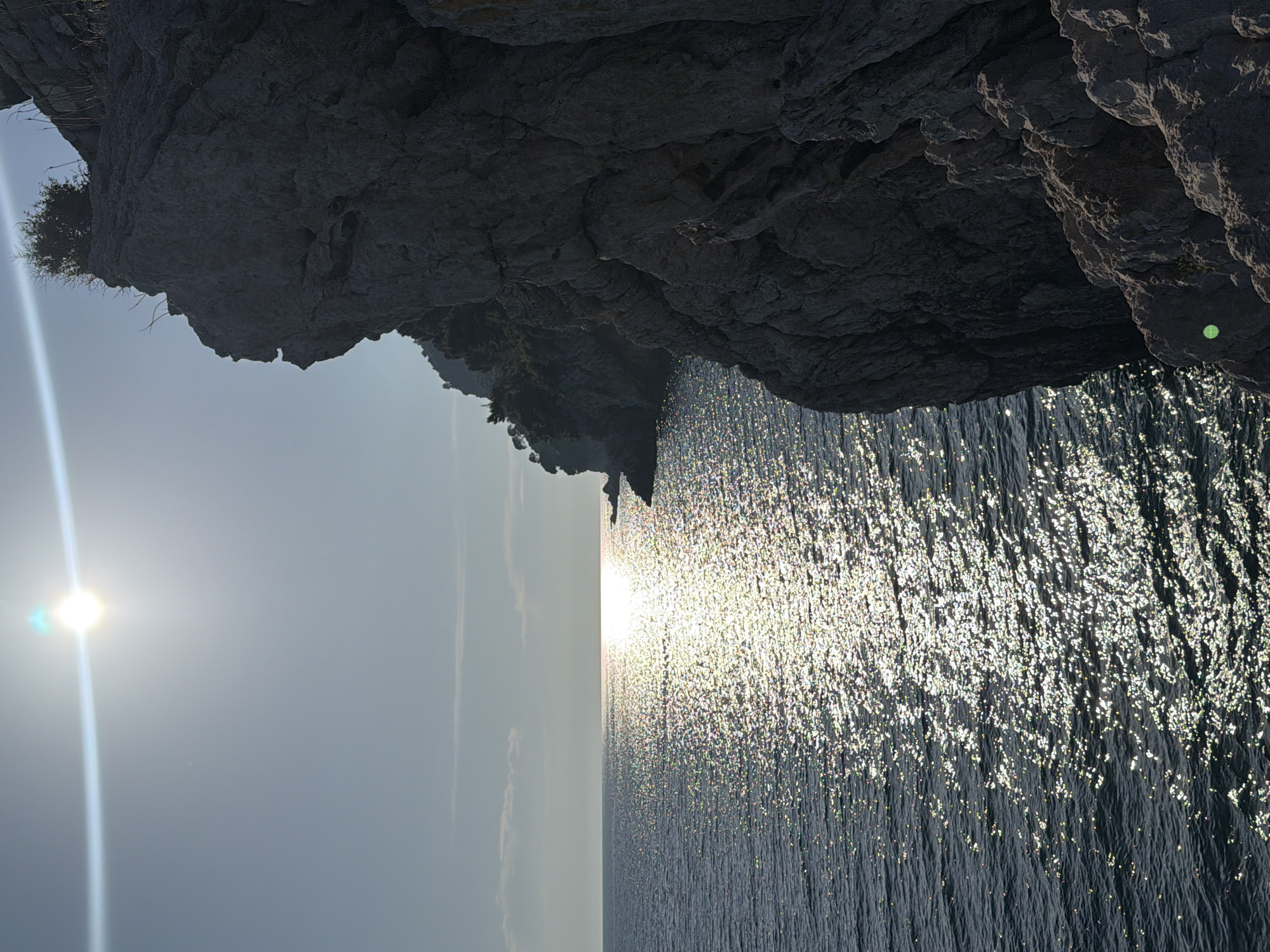 The image captures a sun-drenched seascape with a rocky cliff face in the foreground. The sun is high in the sky, creating a strong lens flare and casting a shimmering reflection across the water. The ocean, a deep blue, is choppy with small waves, and the sun's glare creates a bright path of light extending towards the horizon. The cliff on the right is rugged and grey, with some sparse vegetation clinging to its upper edges. In the distance, along the horizon, a faint outline of land or other cliffs can be seen. The sky is a clear, pale blue, with a few wispy clouds. The scene appears to be a natural, coastal environment, likely a popular spot for scenic views in Petrovac centar, Montenegro.
