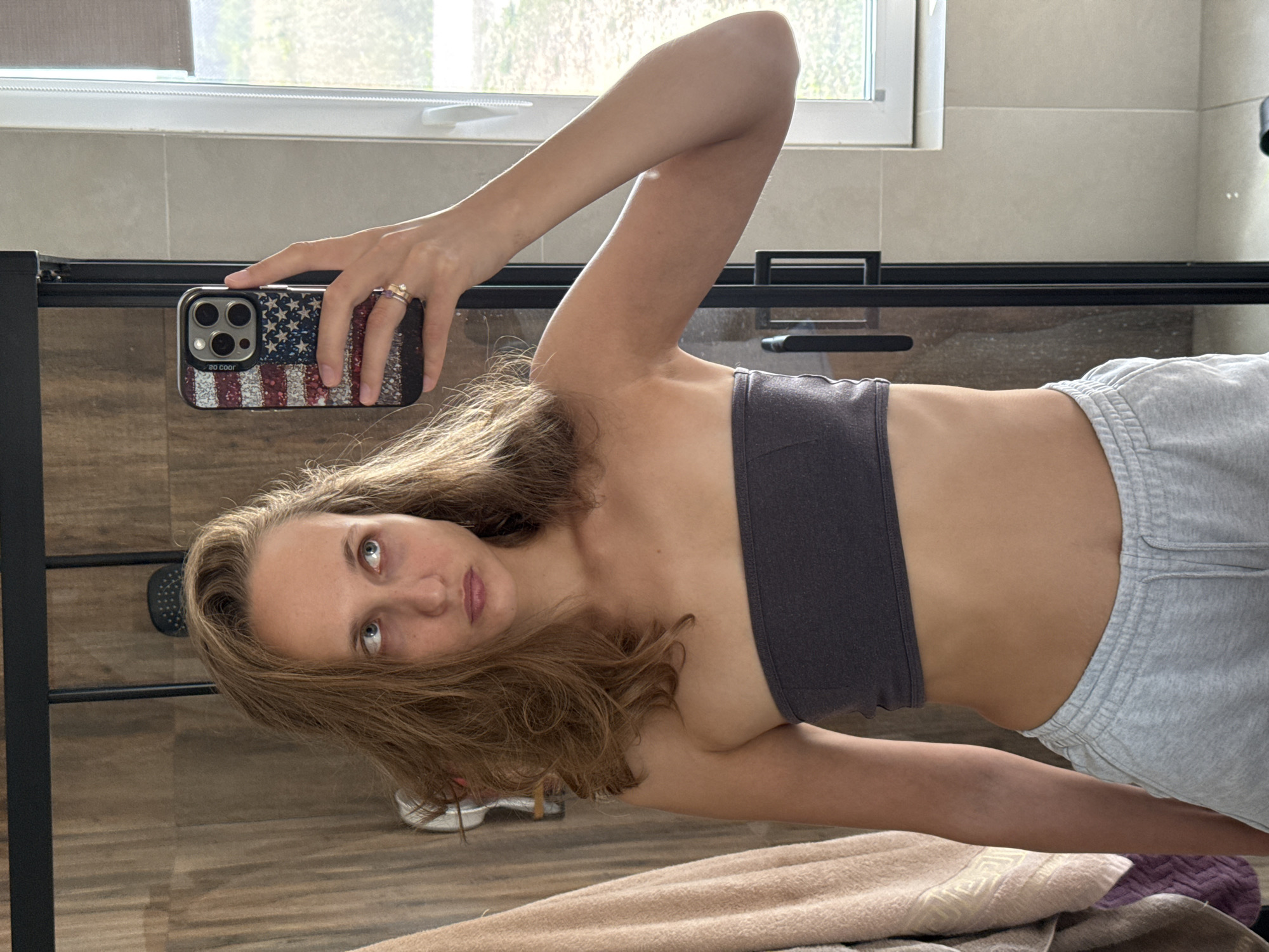 A young Caucasian woman is taking a mirror selfie in a bathroom. She has long, light brown hair and is looking upwards and to her right. She is wearing a dark gray strapless top and light gray sweatpants. Her right arm is raised, holding a smartphone with an American flag-themed case. The background shows a shower with wooden-patterned tiles and a window with blinds. The overall scene appears to be in a private residential bathroom, and the location in the city of Подшивалово, Russia, is not visually indicated in the image.