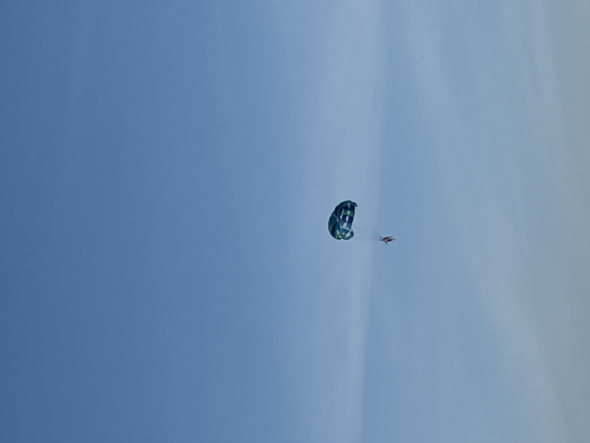 The image shows a person parasailing in a vast, clear blue sky, likely over the ocean given the context of Đà Nẵng, Vietnam. The parasail is predominantly blue and green with a patterned design, catching the sunlight. A small, reddish-brown figure, presumably the person parasailing, is visible suspended below the parachute, attached by ropes. The sky is bright and cloudless, suggesting a sunny day. The overall scene evokes a sense of freedom and exhilaration associated with adventure sports. The lighting indicates it is daytime, likely mid-morning or afternoon given the brightness of the sky. There are no visible text elements in the image.