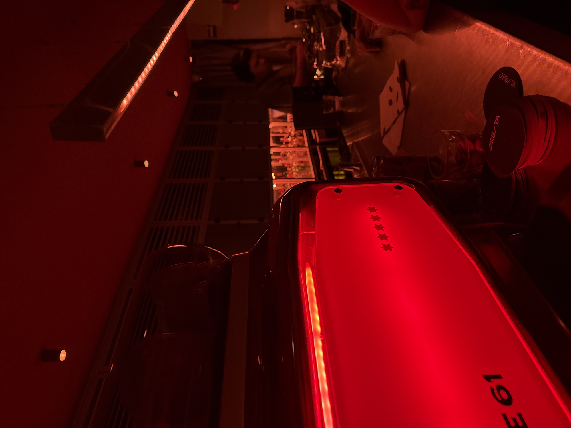 The image depicts a dimly lit bar scene in Mexico City, Mexico, with a strong red color cast. A person, likely a bartender, stands behind a counter with liquor bottles and glasses displayed on shelves. In the foreground, a red-lit appliance, potentially a coffee machine, bears the letters "E 61" and a series of stars. Bottle caps labeled "ORB/TA" sit on the bar top. The environment suggests a modern or trendy establishment.