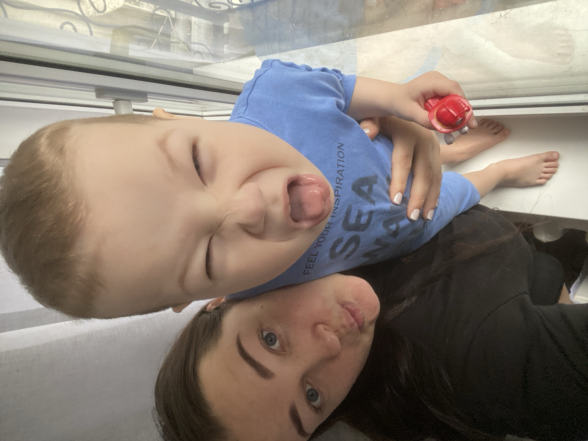 A selfie shows a woman and a child. The woman has dark hair and blue eyes. The child has light hair and is wearing a blue shirt. The child is sticking out their tongue. The woman is lying down. The child is sitting on a window sill and holding a toy. The location of the image is Sochi, Russia.