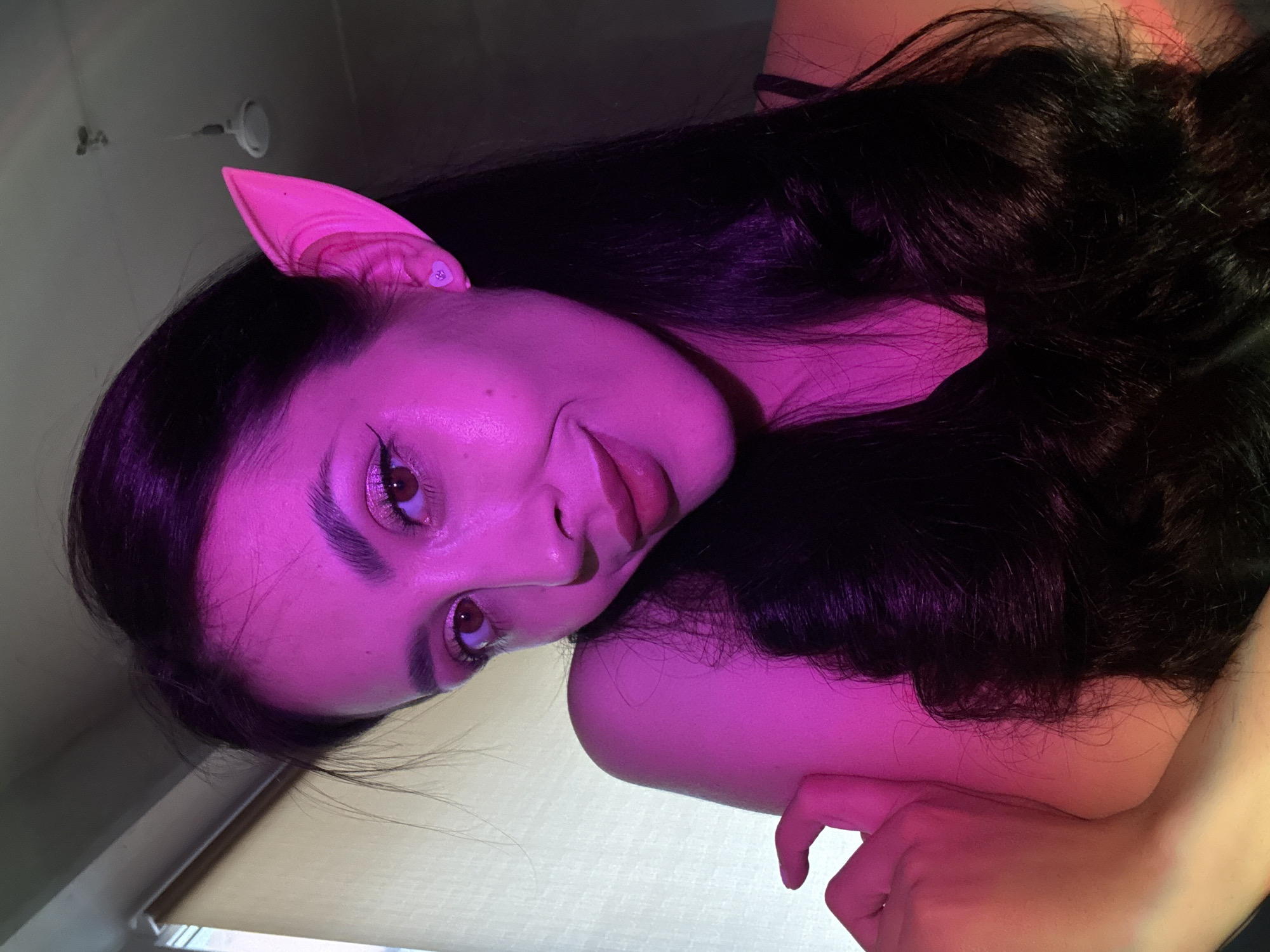The image portrays a person, likely female, with long, dark, wavy hair, illuminated by intense magenta and pink lighting. The person's head is tilted, with their gaze directed towards the viewer, featuring a subtle smirk or knowing expression. They are wearing a prominent, pointed, pink elf-like ear on their right side, adorned with a small, light-colored embellishment near its base.
Their eyes are accentuated with dark eyeliner and possibly shimmery makeup, and their lips are slightly parted. The strong, artificial lighting casts dramatic shadows and highlights across their face and hair, creating a vibrant and somewhat mysterious atmosphere. Part of an arm and hand are visible in the foreground, with fingers gently curled. The background is softly blurred and indistinct, indicating an indoor setting, possibly a wall or ceiling, with faint hints of other ambient light. There is no visible text in the image. The overall impression is that of a posed portrait, possibly for a costume, artistic, or themed photography session.