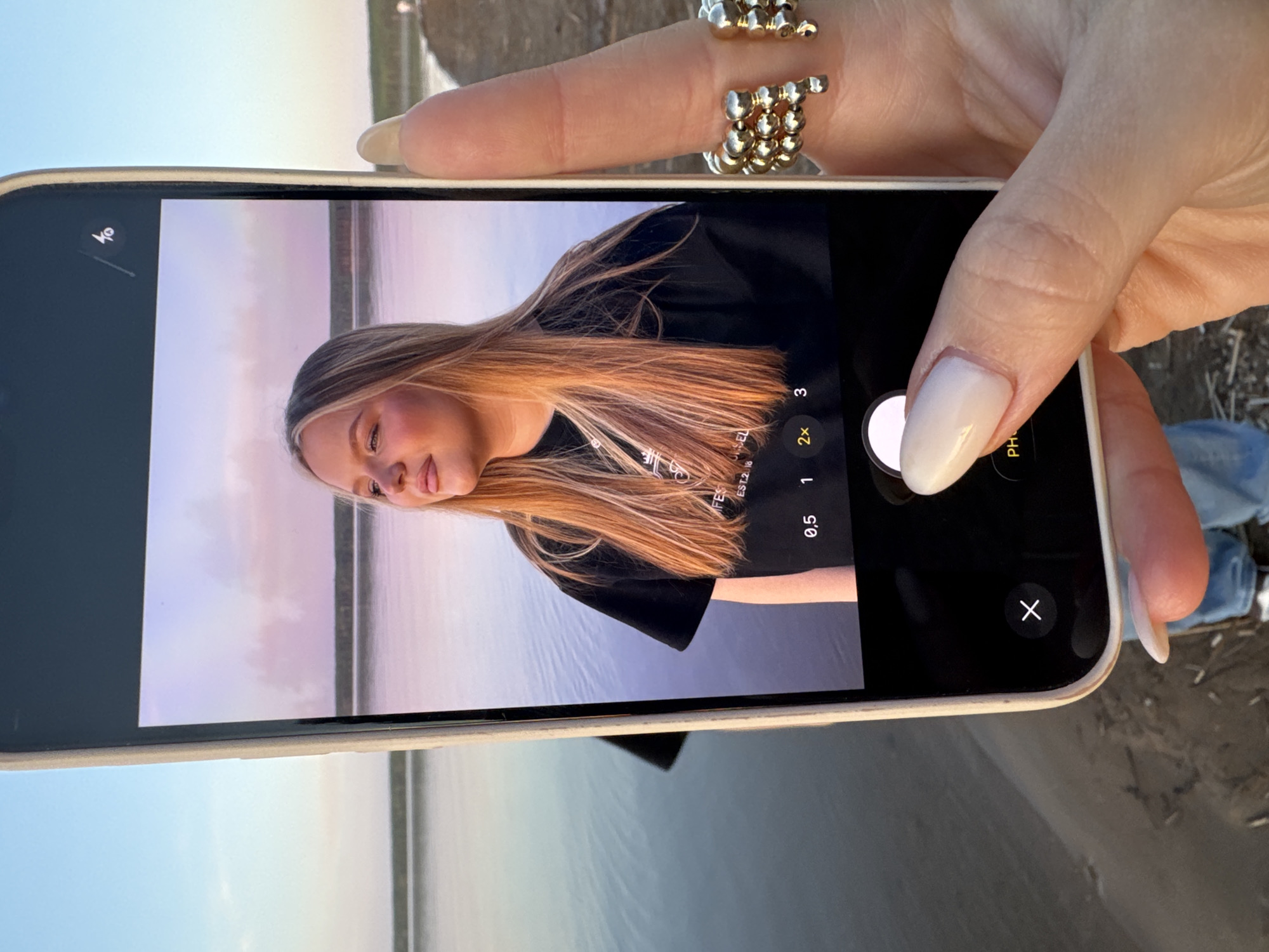 A person's hand is holding a smartphone, displaying a photograph of a woman with blonde hair against a landscape of water and sky with a pinkish hue. The woman in the photograph is wearing a black shirt. The background suggests a shoreline with calm water reflecting the sky. The phone's interface is visible, showing options for zoom and photo settings. The hand holding the phone has polished nails and is adorned with a beaded ring. The surrounding environment appears to be a beach or sandy area, possibly located in the city of Chaikovsky, Russia, if the user's prompt is accurate. The interaction is between the person holding the phone and the photograph displayed on the screen. There is another person out of focus in the background.