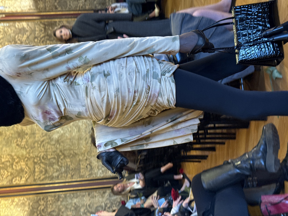 A fashion model walks left-to-right on a polished wooden floor in an ornate, gilded interior. The model is partially visible, wearing a long-sleeved, ruched beige dress adorned with a light pink and green floral pattern, over black opaque legwear. A voluminous draped train extends from the dress's left side. Black leather gloves cover both hands; the model's right hand (viewer's left) clutches a small, glossy black handbag with a crocodile texture. A black, possibly fur, collar or neck covering is visible at the top of the frame. In the background, rows of black chairs suggest audience seating. Visible attendees include a blonde woman in a white top on the mid-left and another person in a dark coat and high-necked garment on the mid-right. The walls feature elaborate golden damask wallpaper or fabric, characteristic of a grand salon or historic building in Paris, France, often used for fashion presentations.