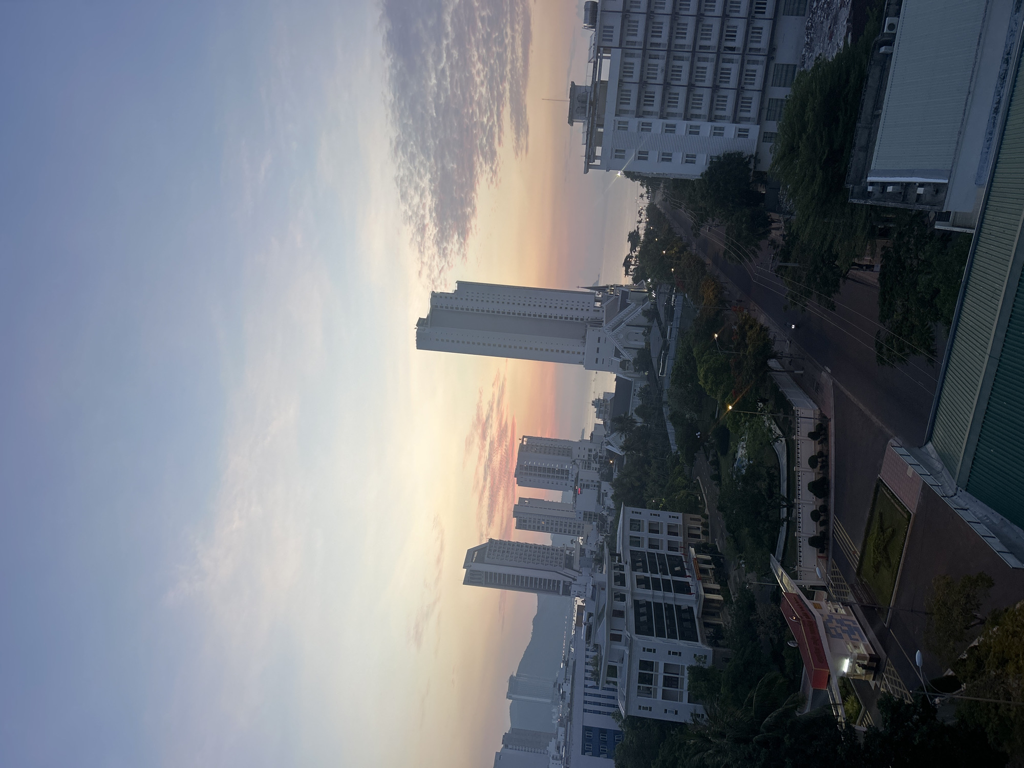 The image captures a cityscape in Bac Nha Trang Ward, Vietnam, at dawn or dusk. The sky transitions from a light blue to shades of pink and orange near the horizon. Several tall, modern buildings with repeating window patterns dominate the urban landscape. A road lined with trees runs through the center of the view. The architectural styles suggest a mix of residential and commercial structures. Some vegetation is visible among the buildings.