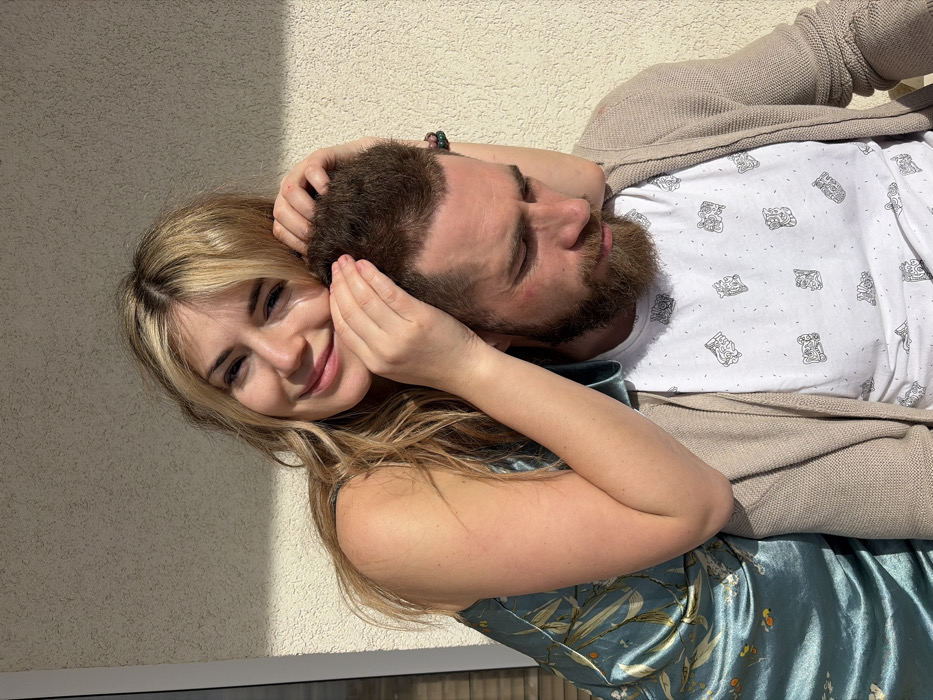 A close-up photograph captures two individuals outdoors in Budva, Montenegro, illuminated by direct sunlight against a textured, light-colored stucco wall. A female, appearing to be in her 20s to 30s, is positioned behind and to the left of a male. She has long, wavy blonde hair, a fair complexion, and is smiling directly towards the camera. She wears a sleeveless, light blue satin-like dress with a subtle botanical pattern. Her hands are placed on the top of the male's head, with her right arm extended around his upper back/shoulder. The male, appearing to be in his 30s to 40s, has short brown hair and a full reddish-brown beard. He is looking downwards, away from the camera. He wears a white t-shirt with scattered black graphic symbols and a light beige knitted cardigan. A dark beaded bracelet is visible on his left wrist. Strong shadows are evident on the subjects and the background wall.