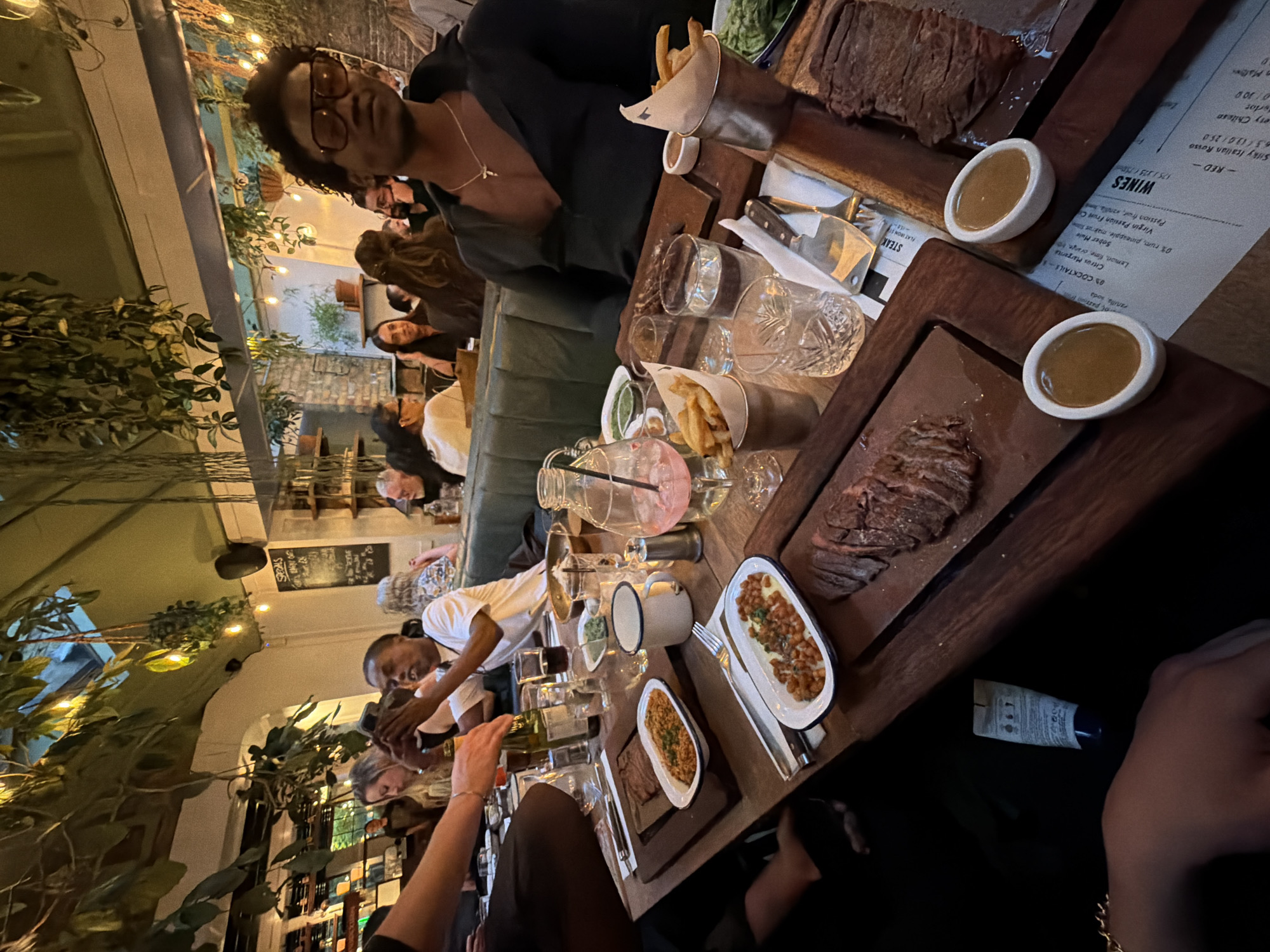 This image captures a lively and atmospheric dining scene inside a restaurant or bar, likely during the evening given the warm, artificial lighting. The perspective is rotated 90 degrees clockwise, placing the table and diners vertically in the frame.
In the foreground, a person with dark, curly hair and glasses, wearing a dark top with a gold necklace, looks towards the camera. They are seated at a wooden table laden with food and drinks. Two large wooden boards prominently display sliced steak, accompanied by small white bowls of a brown sauce and paper cones filled with french fries. Other dishes on the table include white oval plates containing what appears to be beans and rice. Various clear glasses, some empty and some containing drinks, along with a large glass pitcher holding a pink beverage, are also visible. Another person in a white shirt is seen interacting with a bottle, possibly pouring a drink.
The setting features abundant green foliage and string lights draped overhead, creating a cozy, garden-like ambiance. In the background, numerous other patrons are seated at tables and along a green banquette, engaged in conversation and enjoying their meals. The walls appear to be light-colored brick or plaster.
Visible text on menus includes sections for "WINES" (e.g., "Silky Italian Rosso," "Chilean," with prices like "6.5 / 13.0 / 25.0" and "17.5 / 32.5 / 75.0"), "COCKTAILS" (e.g., "Citrus Margarita," "Sober Mule," "Virgin Passion Fruit Cooler," listing ingredients like "Lemon, lime, orange" and "0% rum, pineapple, makrus"), and "STEAK" (specifically "FLAT IRON ST"). A sign with "SPROMS" is also partially discernible.
The overall atmosphere suggests a casual and social dining experience in what could be a popular spot in London, United Kingdom.