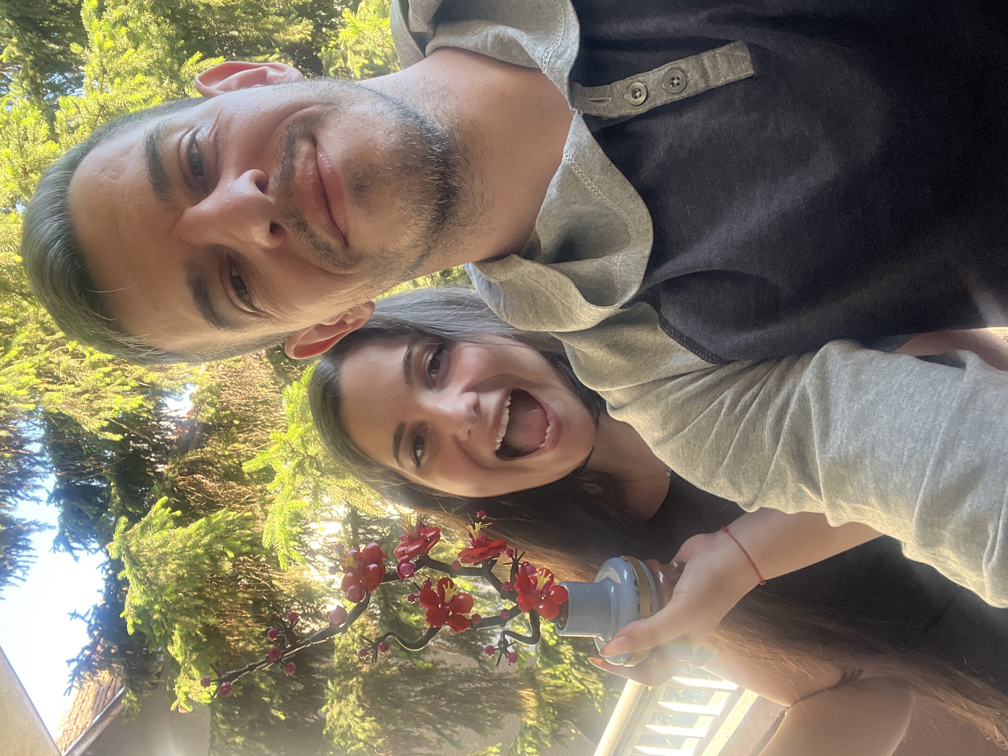 A Caucasian man and woman are taking a selfie outdoors, with a bright green coniferous tree and a building roof in the background. The man, in the foreground, is looking at the camera with a slight smile and stubble. He is wearing a dark grey and light grey raglan-style shirt with buttons at the collar. The woman is behind him, holding a small decorative vase with red flowers, and smiling widely with her mouth open. She has long, dark brown hair and is wearing a dark top. A thin red bracelet is visible on her wrist. The scene appears to be in a residential area, possibly a garden or balcony, with a white railing visible in the lower left corner. The lighting suggests it is daytime, with sunlight filtering through the trees. The image was taken on July 23, 2023, at 17:12, indicating a late afternoon setting. It is plausible this location is in Sofia, Bulgaria, given the context of other images from the same source.