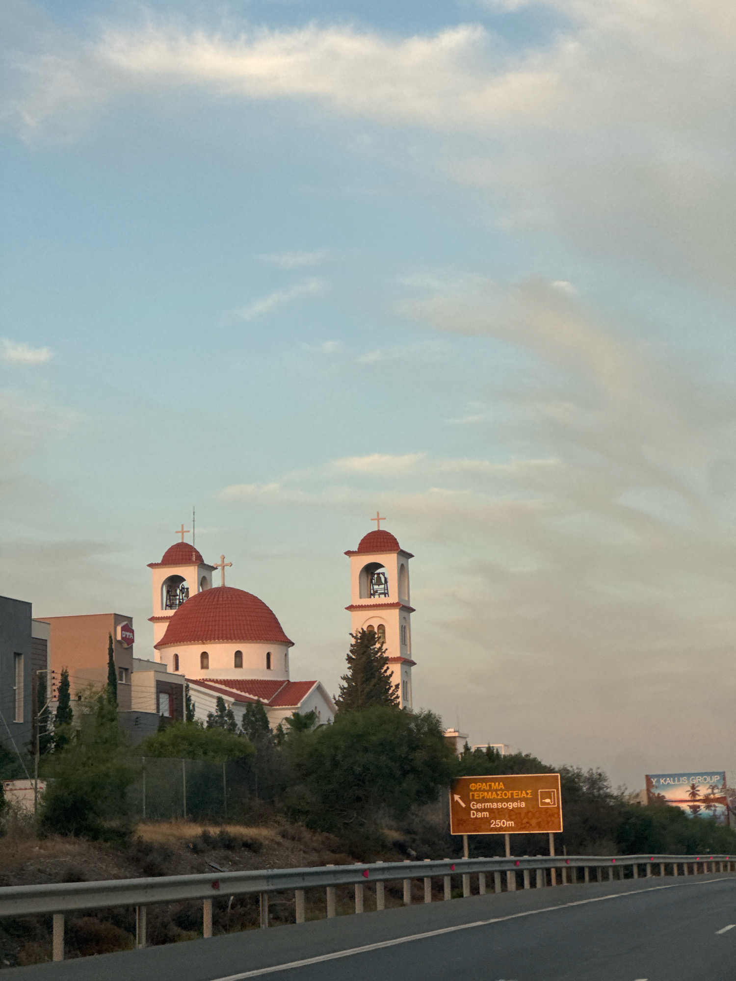 The image shows a road scene in Germasogeia Municipality, Cyprus. A white church with red tiled domes and bell towers is visible in the background. A road sign indicating "FRAGMA GERMASOGEIA Dam 250m" is also present, along with a billboard for "Y. KALLIS GROUP". The sky is partly cloudy and the scene appears to be captured during daylight hours.