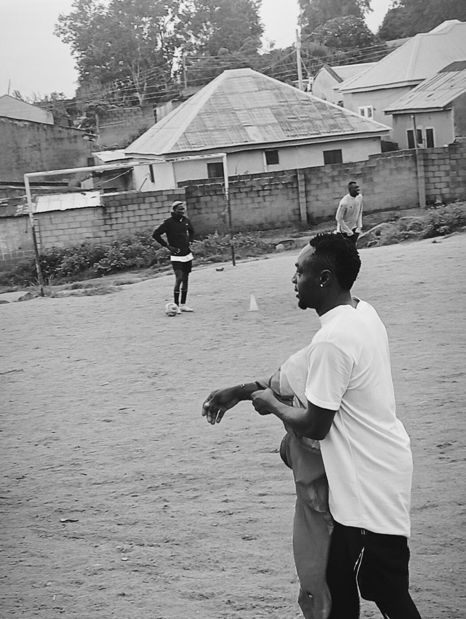In Jos, Nigeria, a black and white image depicts a sandy field, likely a makeshift soccer pitch, with a few scattered players. In the foreground, a man wearing a white t-shirt and black shorts with white stripes faces away from the camera, his profile visible. He appears to be interacting with another person, whose hands and arms are the only visible part of their body, also in the foreground. In the middle ground, another man stands with hands on his hips, wearing a dark jacket, white t-shirt, and shorts, with a soccer ball at his feet. Further in the background, a third man in a light-colored shirt walks away from the camera. Behind the players, a low brick wall separates the field from a street lined with buildings, some with corrugated metal roofs, and lush green trees. A soccer goal is partially visible on the left side of the frame. The overall impression is of an informal sporting activity taking place in a casual urban environment.