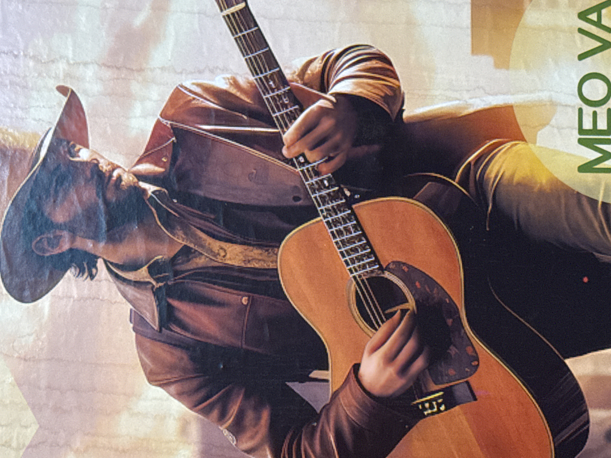 A man wearing a brown leather jacket and a cowboy hat plays an acoustic guitar. Part of the name "MEO VA" is visible in green on the right edge of the image. The location is likely Mèo Vạc Commune, Vietnam, based on the text fragment.