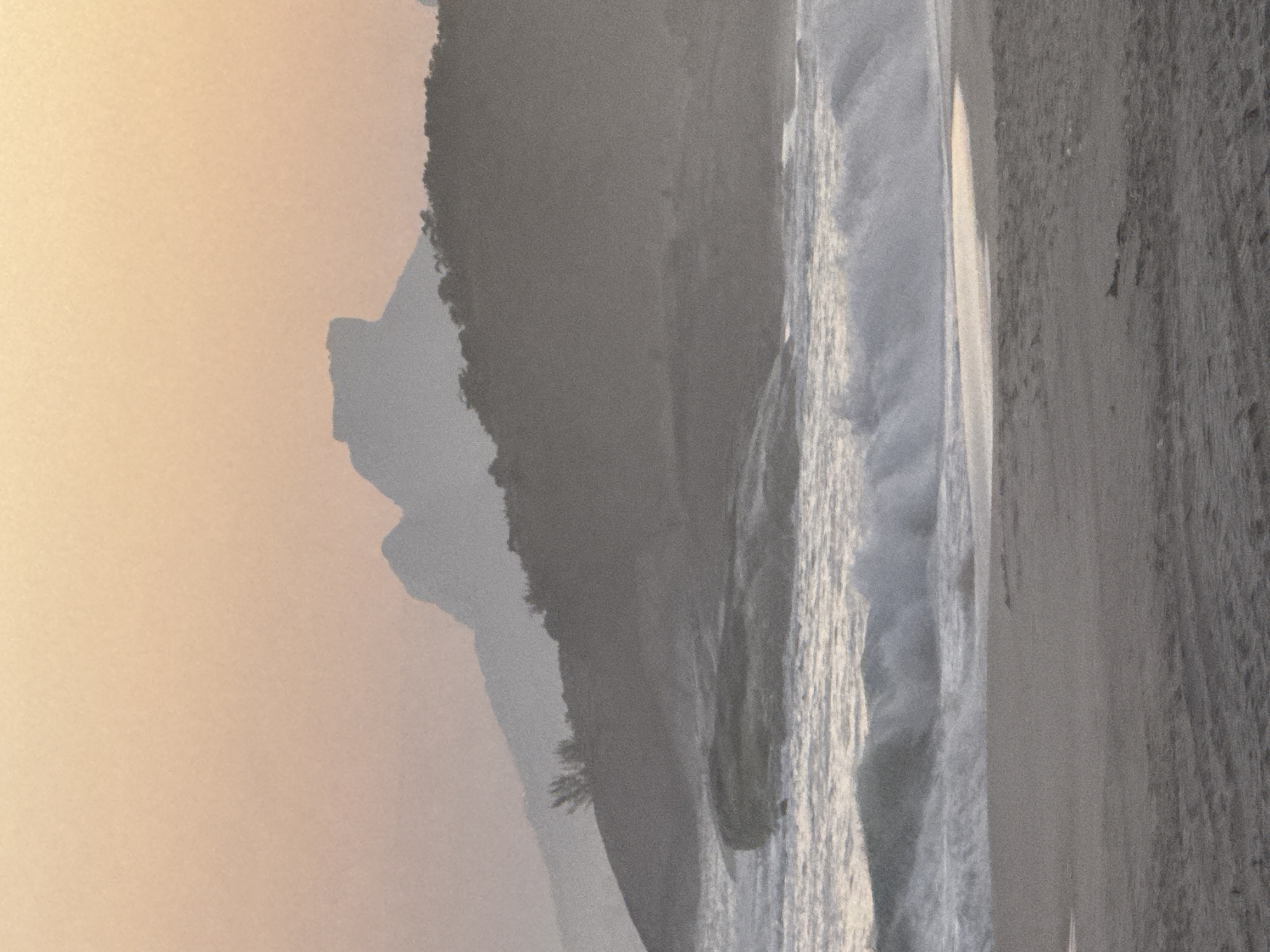 The image depicts a serene beach scene at dusk, likely in Niterói, Brazil, given the iconic silhouette of the mountains in the background. The sky is a soft gradient of peach and orange, transitioning to a hazy, muted gray as it meets the distant mountains. These mountains, rendered in silhouette, are the most prominent feature of the background, with their rugged shapes clearly defined against the sky.
In the midground, a lush, tree-covered hillside slopes down towards the water. Waves are breaking on the shore, creating a foamy white line that contrasts with the darker sand. The water itself appears choppy, with visible whitecaps and the movement of the surf. Large, dark rocks are scattered along the shoreline, some partially submerged by the waves.
The foreground consists of a sandy beach, appearing slightly damp near the water's edge. The sand has a textured appearance, with subtle variations in color and the presence of small footprints or imprints. The overall impression is one of natural beauty and tranquility, enhanced by the soft, diffused light of sunset. No people or animals are visible in the frame.