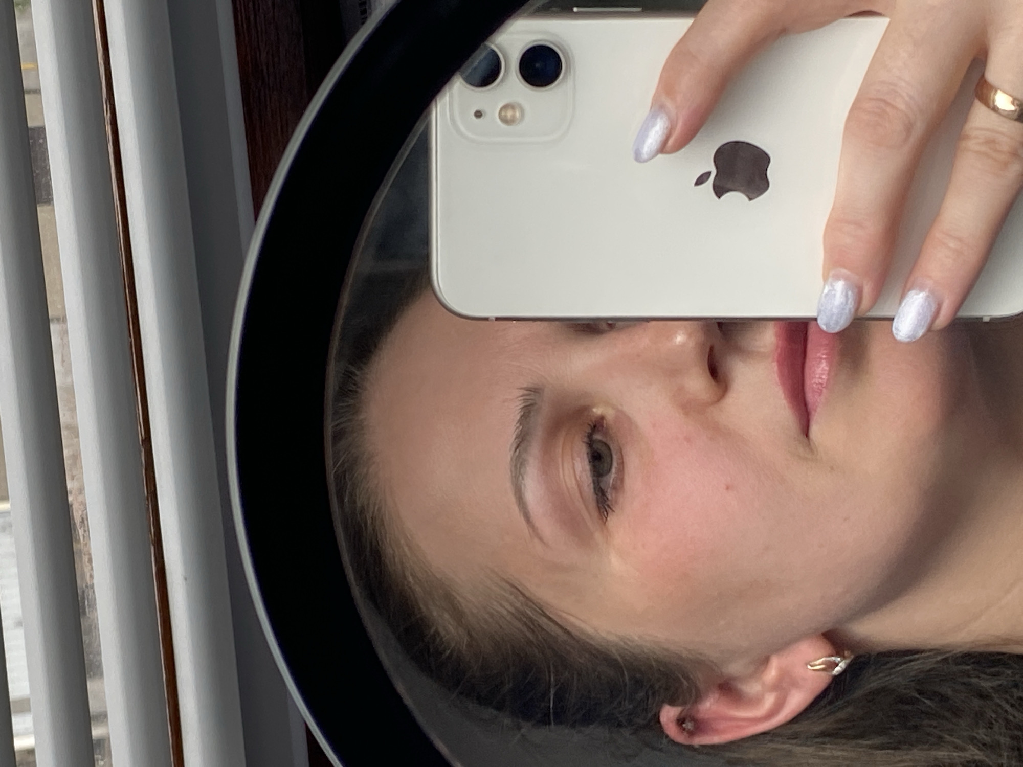 A woman's face is partially visible in a mirror selfie taken with a white iPhone. The woman is holding the phone so that the camera section covers the bridge of her nose. Her left eye is visible and a portion of her lips. She has manicured nails with a light color and a ring on her right hand. The woman's left ear is visible, featuring an ear piercing. The background includes a window with white blinds and a dark wooden frame around the mirror. The location is not discernable.