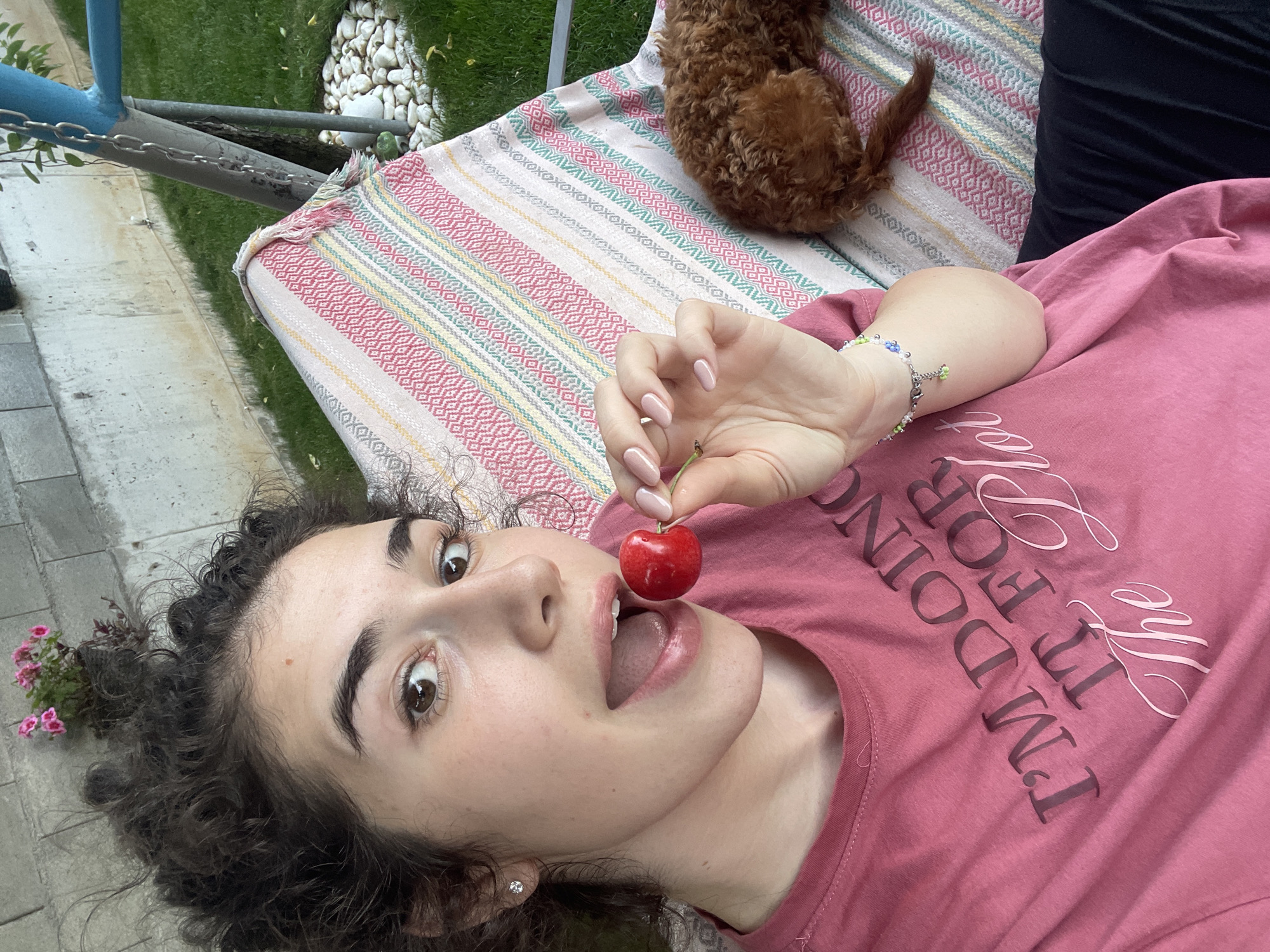 A woman with curly dark hair lies on a striped textile, holding a red cherry at her mouth. She wears a pink t-shirt with a printed phrase on it and a bracelet on her left wrist. Part of a brown dog rests on the textile next to her. The scene appears to be outdoors, with grass and white stones visible in the background, along with part of a blue metal structure. The location is likely in a residential area. There is no indication in the photograph that indicates the city of Sofia, Bulgaria.
