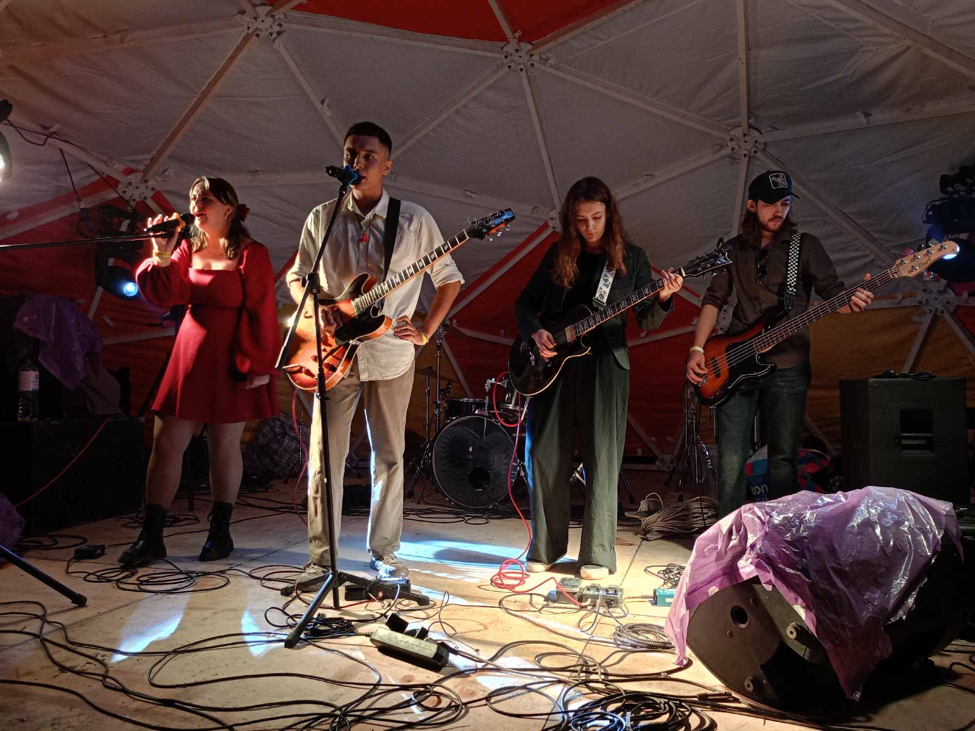 A band is performing on a stage inside a geodesic dome structure. Four individuals are present: a female singer in a red dress, a male guitarist in a white shirt, a female guitarist in a dark jacket, and a male bassist wearing a cap. The stage is covered in black cables and appears to be made of a concrete or stone surface. A drum set is visible behind the guitarists. Speakers are set up on the stage, one of which is covered with purple plastic sheeting. The lighting is dim, with spotlights illuminating the performers. The overall atmosphere suggests an indoor concert or performance. The geodesic dome structure suggests a temporary or open-air venue. The scene does not provide any specific visual cues to identify the location as Barnaul, Russia, or any other specific city.
