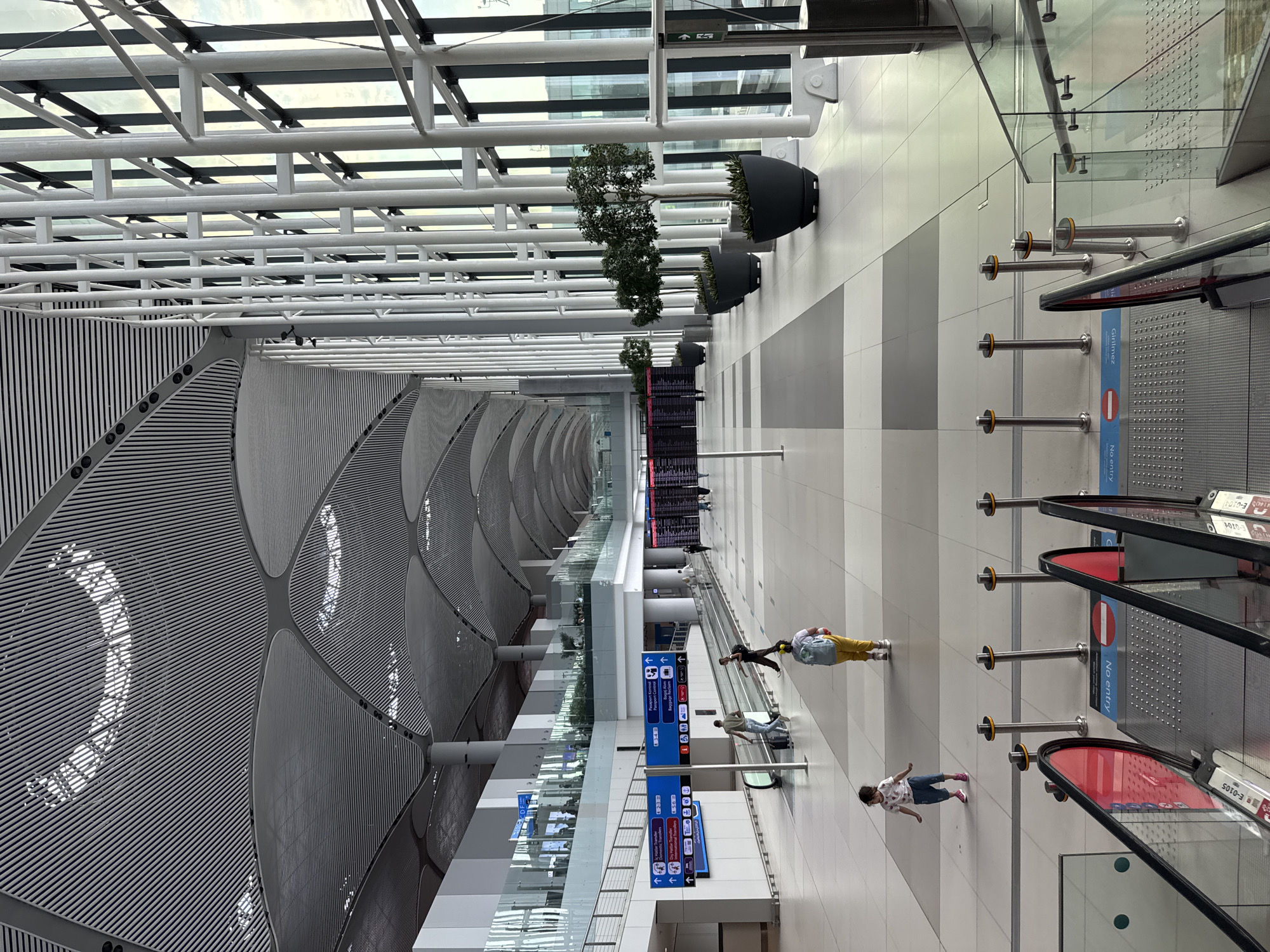 The image shows a spacious, modern airport terminal with a high, vaulted ceiling supported by numerous white, structural pillars. Large windows line one side of the terminal, allowing natural light to illuminate the interior. The floor is a tiled expanse of white and gray. On the left side of the terminal, there are multiple digital display boards showing flight information. Several individuals are walking through the terminal, including a child in a white shirt and blue pants, and another person in yellow pants and a blue backpack. Escalators and moving walkways are visible, with people using them. Potted trees are placed along the side of the terminal near the windows. The location is identified as Arnavutköy, Turkey, likely within the Istanbul Airport.