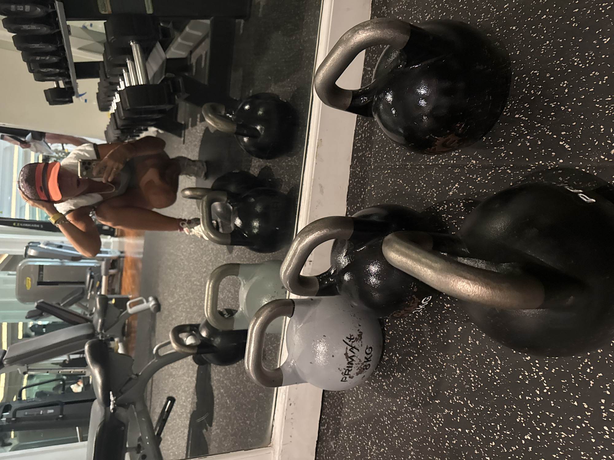 A mirrored gym scene features a woman in a squat taking a selfie with a smartphone, with rows of dumbbells visible in the mirror’s reflection. The foreground highlights kettlebells of varying sizes and colors on a speckled black floor. The gym setting includes various exercise machines and equipment in the background. The location is Kuta Selatan, Indonesia.