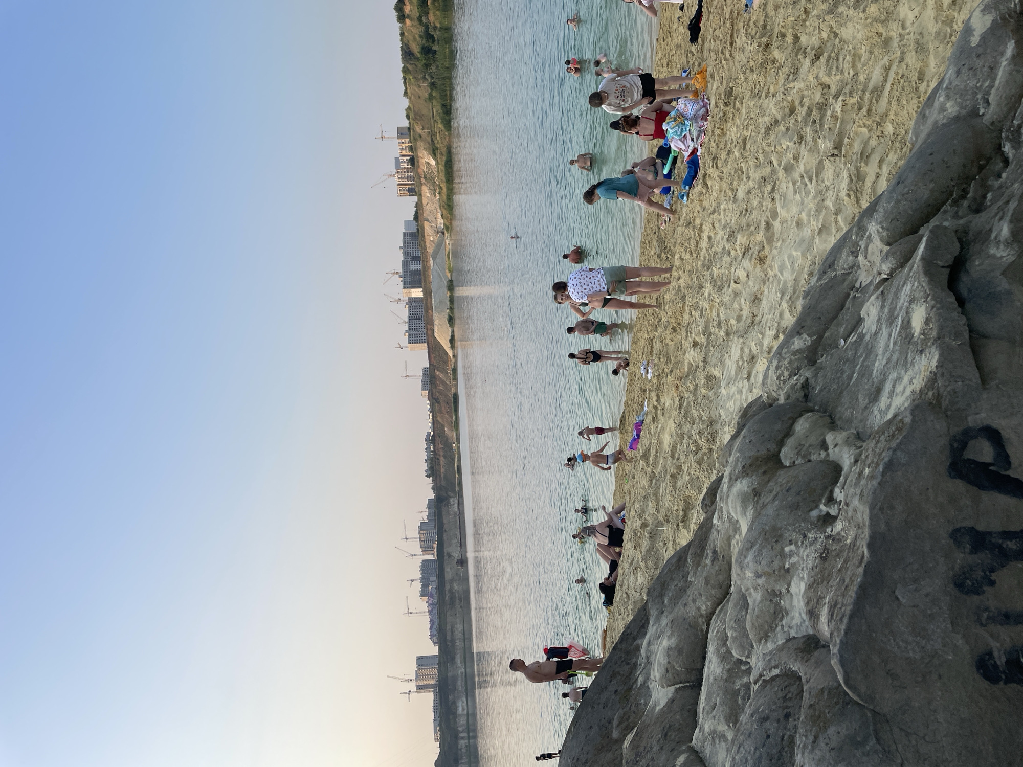 The image depicts a quarry lake in Tambov, Russia, on a clear day with calm water and a sandy beach. Numerous people are present, engaged in various activities. Several individuals are swimming or wading in the shallow parts of the lake, while others are relaxing on the sand. Some are sunbathing, sitting on towels or directly on the sand. A group of people are gathered on the beach, some interacting with each other. In the background, a constructed embankment with buildings and cranes suggests ongoing development. The foreground is dominated by large, textured rocks and the sandy beach. The sky is clear and blue, fading to a lighter shade near the horizon.