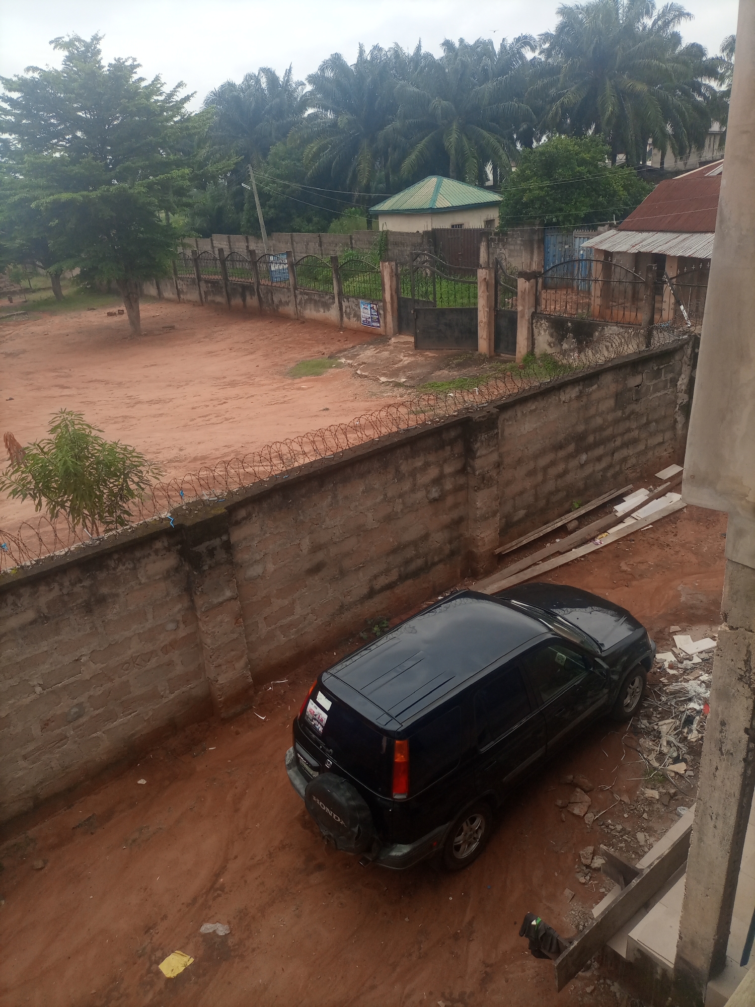 This media file captures an outdoor scene in Nkpor, Nigeria, under an overcast sky, suggesting a cloudy or potentially rainy day. The primary subjects are a black Honda CR-V parked on a dirt lot, and a weathered brick wall topped with barbed wire.
The setting appears to be a somewhat unkempt urban or semi-urban area, with a mix of residential and possibly commercial elements visible in the background. Palm trees and other foliage are abundant, indicating a tropical environment. Behind the wall, a fence with decorative metalwork separates the property from the street. In the distance, various buildings can be seen, including one with a green, pitched roof and another with a rusty, corrugated metal roof. The ground is predominantly red dirt, with some scattered debris and a few small plants growing near the wall.
There are no people or specific activities actively taking place in the image. The focus is on the static elements of the scene: the car, the wall, the vegetation, and the buildings. Notable details include the spare tire mounted on the back of the Honda CR-V, which has the "HONDA" brand clearly visible. The car also has several stickers on its rear window, though their content is not discernible. The brick wall shows signs of age and weathering, with some moss or discoloration.
The overall impression is one of a typical day in a Nigerian town, with a focus on the physical environment rather than human interaction. The cloudy weather contributes to a subdued atmosphere. No specific text is clearly legible on the buildings or fences, except for what appears to be signage on posters attached to the fence near the entrance, and the branding on the car.