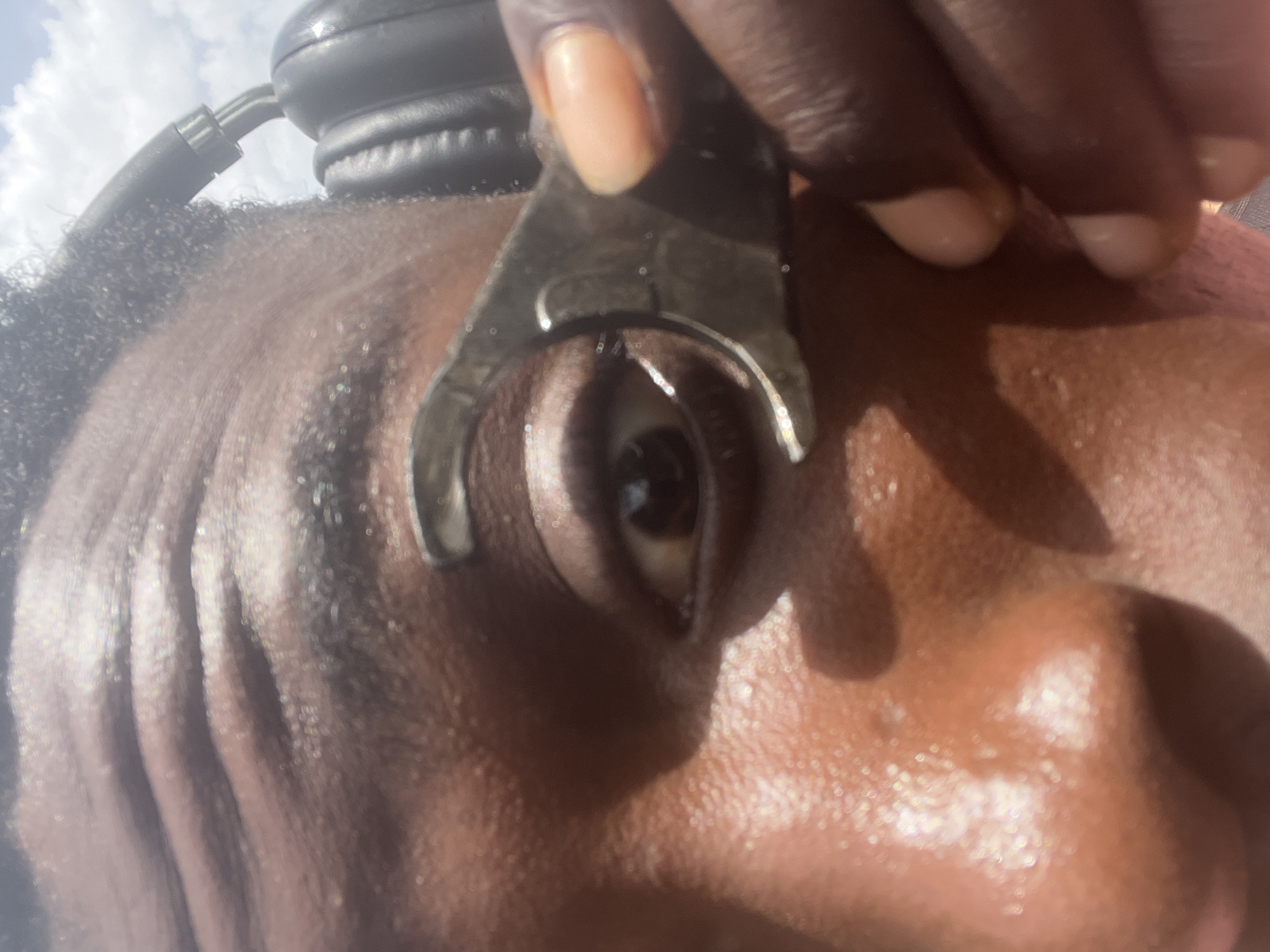 A close-up, high-angle shot captures the upper right side of a person's face, focusing on the eye, forehead, and part of the cheek. The individual has dark brown skin and short, dark hair. Black over-ear headphones are visible on the head, partially obscured at the top. A hand with dark skin and light-colored fingernails is holding a metallic, C-shaped tool, possibly an eyelid speculum, which is carefully positioned to hold the person's right eye open. The eye's pupil and iris are clearly visible, appearing to look slightly upwards. The skin on the forehead and cheek has a noticeable sheen, reflecting bright light, which may suggest sweat or oil, and indicates outdoor conditions. In the blurred background, a bright, hazy sky with visible white clouds confirms an outdoor setting during daytime. The overall impression is an eye examination or procedure. No text is visible in the image. The event takes place in Nkpor, Nigeria.