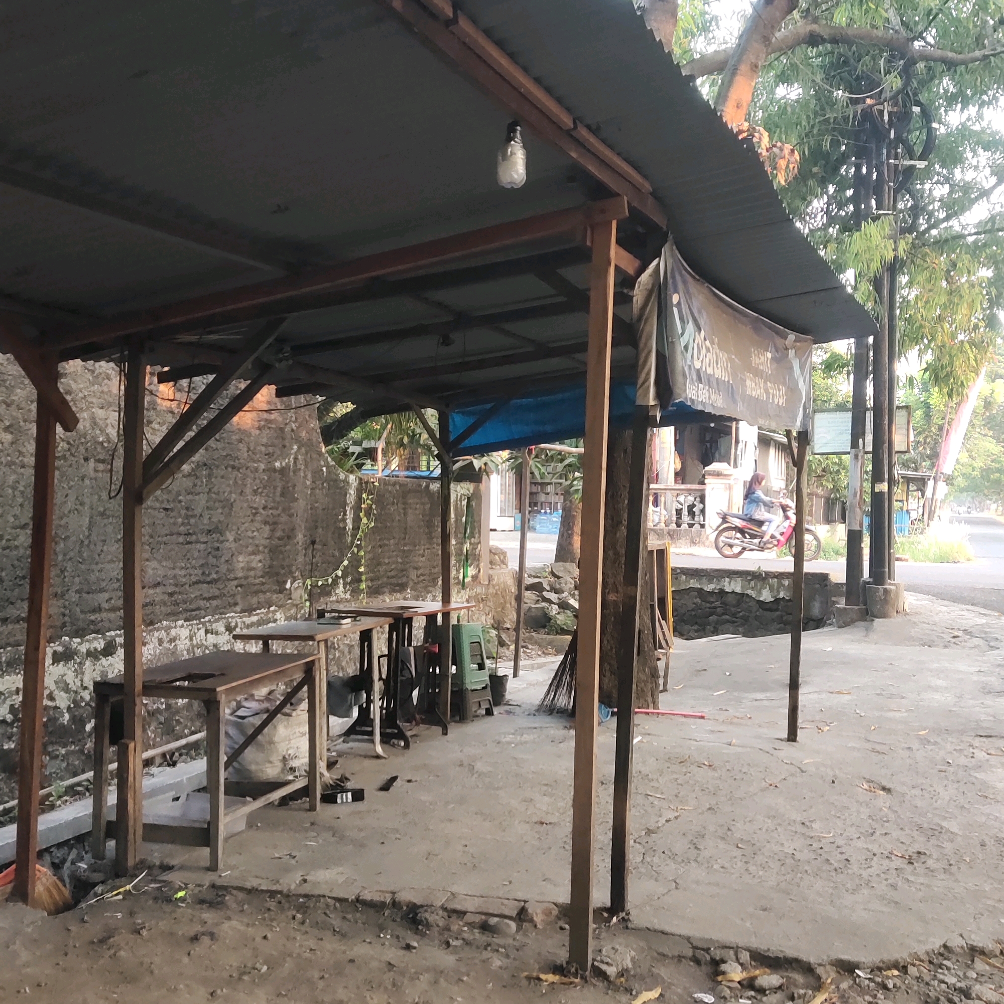 The image depicts an outdoor scene in Sragen, Indonesia, likely on the side of a road. In the foreground, there is a covered area with a corrugated metal roof supported by wooden posts. Beneath the shelter, several wooden tables and chairs are arranged on a concrete floor. A green plastic chair is visible, along with a broom leaning against a wooden post. The background shows a textured stone wall, some vegetation, and a road with a woman riding a pink motorcycle. A banner with text, possibly advertising a business, hangs from the roof. The environment appears somewhat unkempt, with debris on the ground. The lighting suggests it is either early morning or late afternoon.