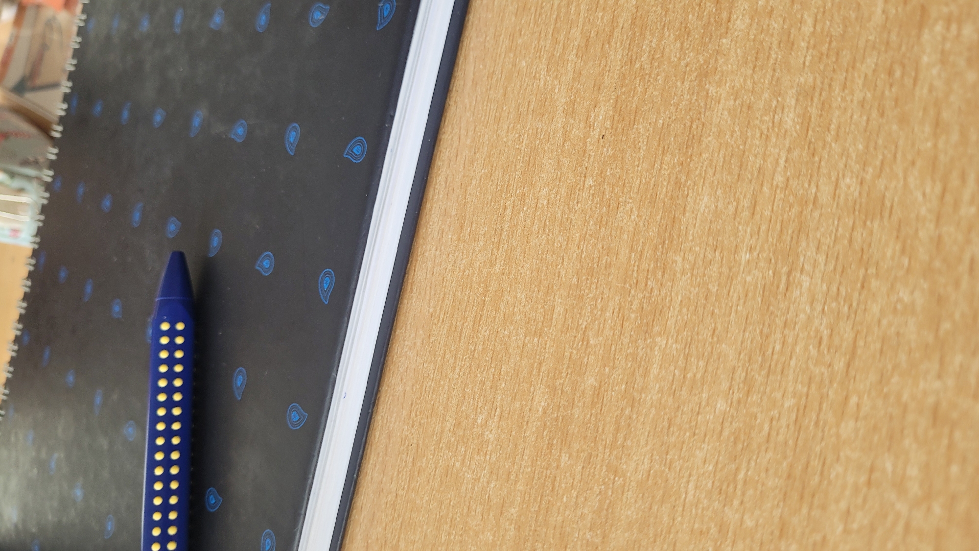 The image shows a dark blue pen with yellow dots lying on a black notebook with a repeating paisley pattern in a lighter blue. The notebook sits on a surface that appears to be light brown wood. The pen is near the edge of the notebook. The image appears to be taken indoors. There is no specific information in the image to suggest it was taken in Varna, Bulgaria.