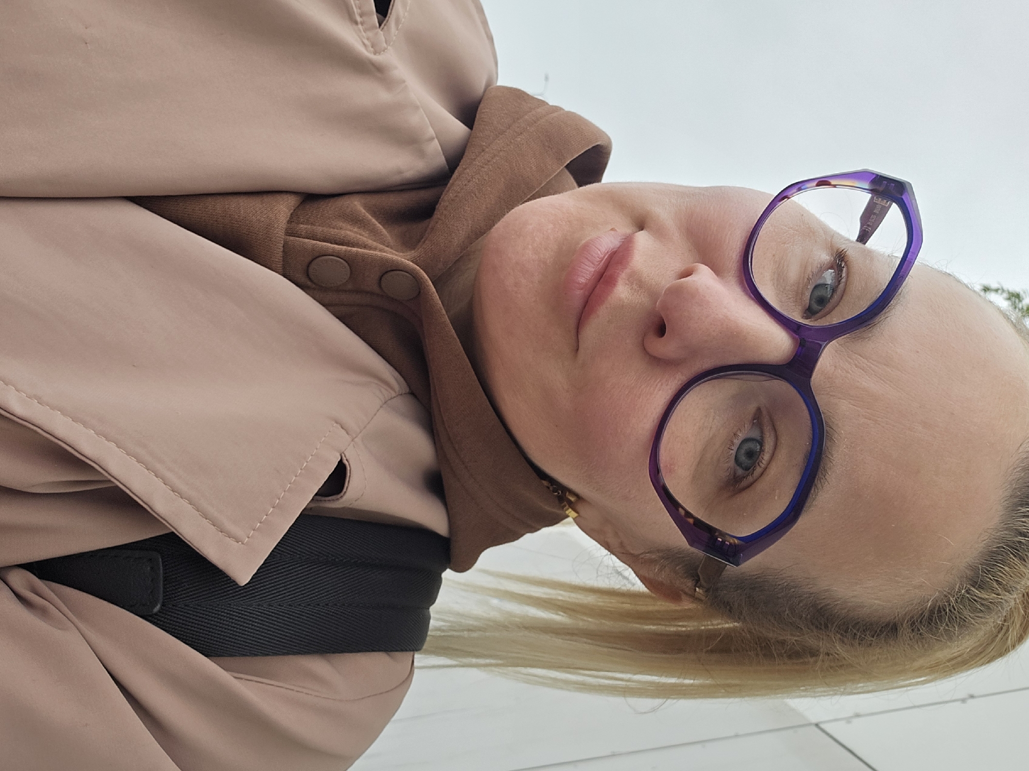 A woman with fair skin and blonde hair poses in a tilted portrait. She wears purple glasses and a light pink trench coat over a brown top with buttons. A black strap is visible under the coat. The background appears to be a light gray surface, possibly an outdoor tile. There is no definitive information in the image to confirm the location is Sochi, Russia.